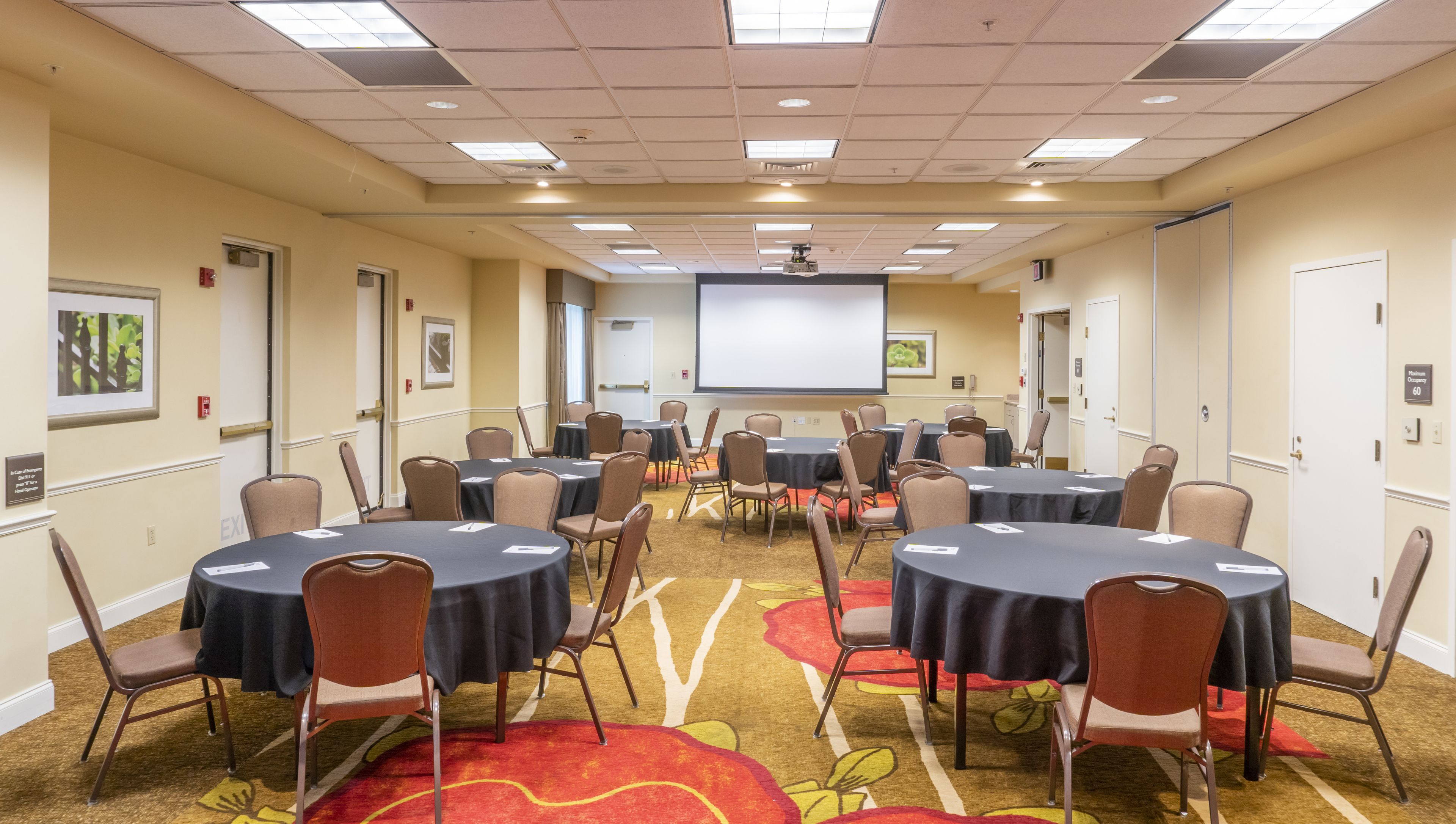 A conference room with multiple tables and chairs, a projector screen, and a carpeted floor.