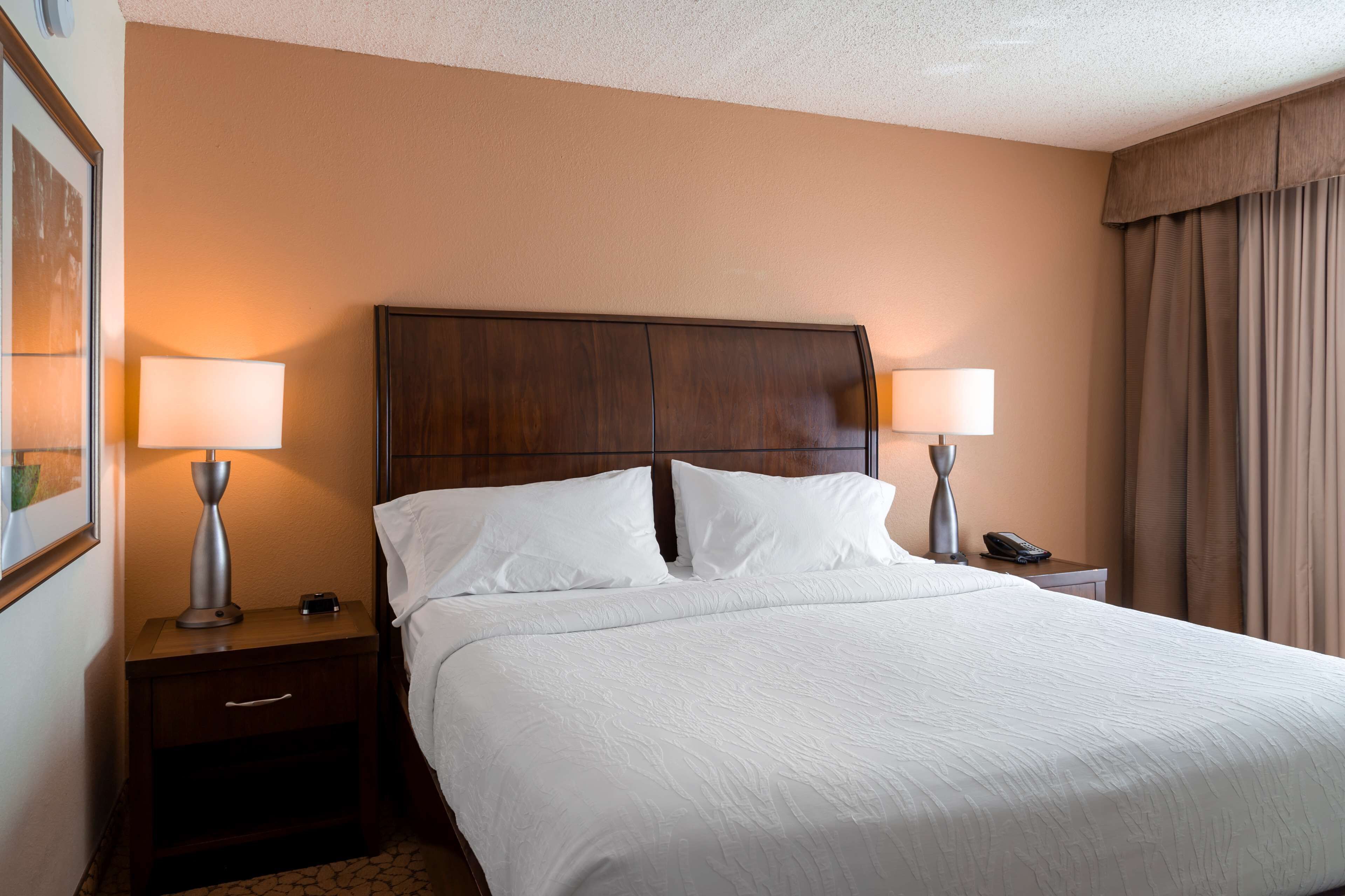 A hotel room with a large bed, two bedside lamps, a wooden headboard, a nightstand with a phone, and a framed picture on the wall.