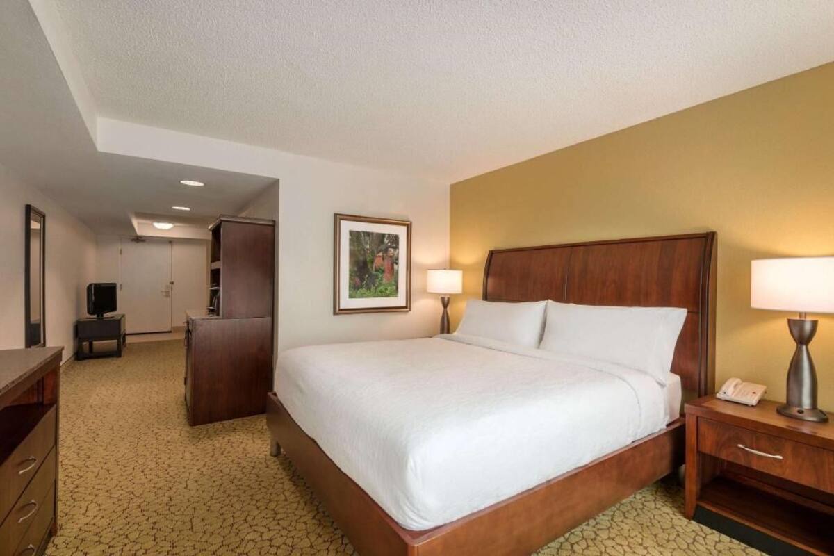 A hotel room with a bed, bedside lamps, a desk, a chair, a television, and a framed picture on the wall.