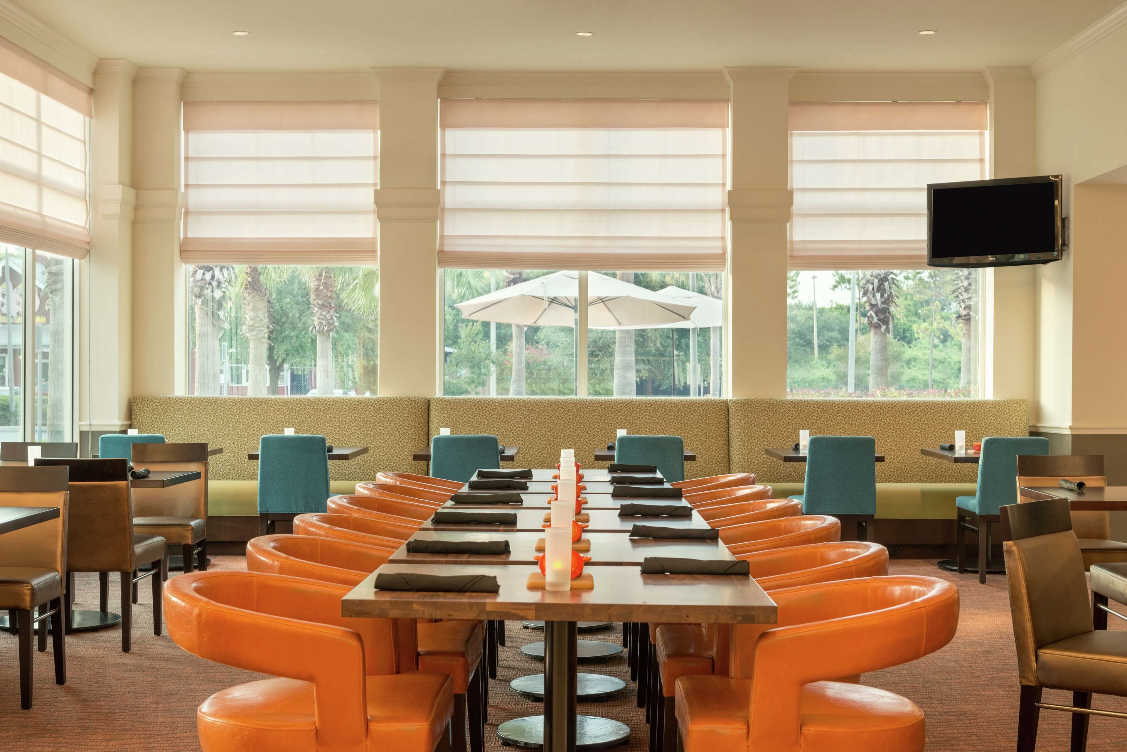 A dining area with long tables and orange chairs, large windows with blinds, and a flat-screen TV on the wall.