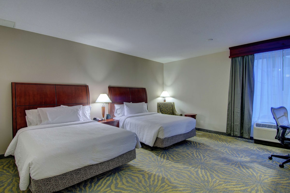 A hotel room with two beds, a desk with a lamp, a chair, and a large window with curtains.