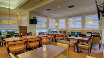 Un comedor amplio con varias mesas y sillas, ventanas grandes con persianas e iluminación colgante.