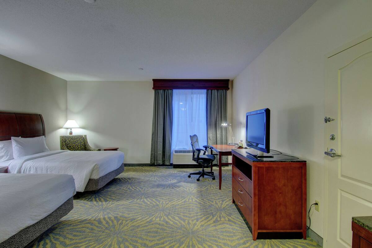A hotel room with two beds, a desk with a computer, a television, and a window with curtains.