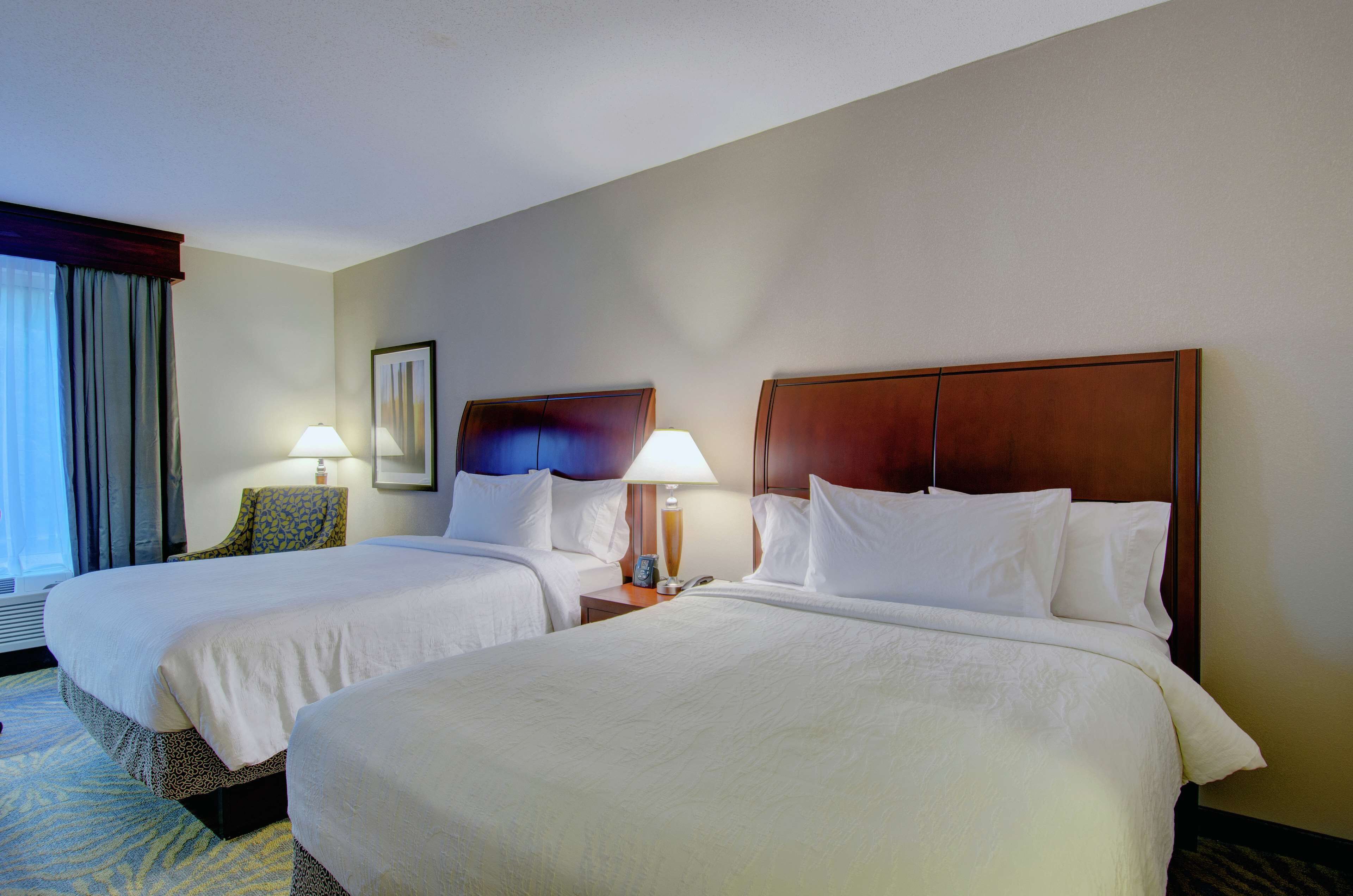 A hotel room with two beds, a nightstand with a lamp, a framed picture on the wall, and a window with curtains.