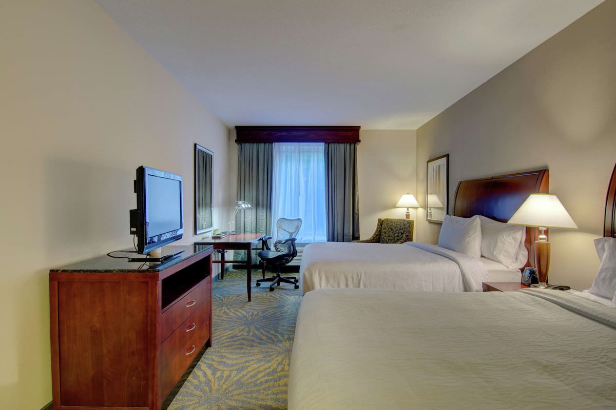 A hotel room with two beds, a desk with a chair, a television, and a window with curtains.