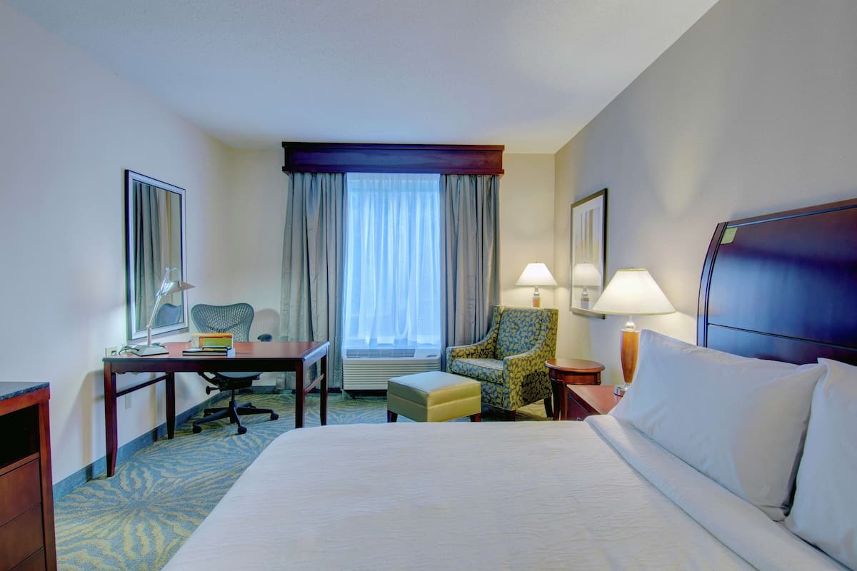 A hotel room with a large bed, a desk, a chair, and a small table with a lamp.