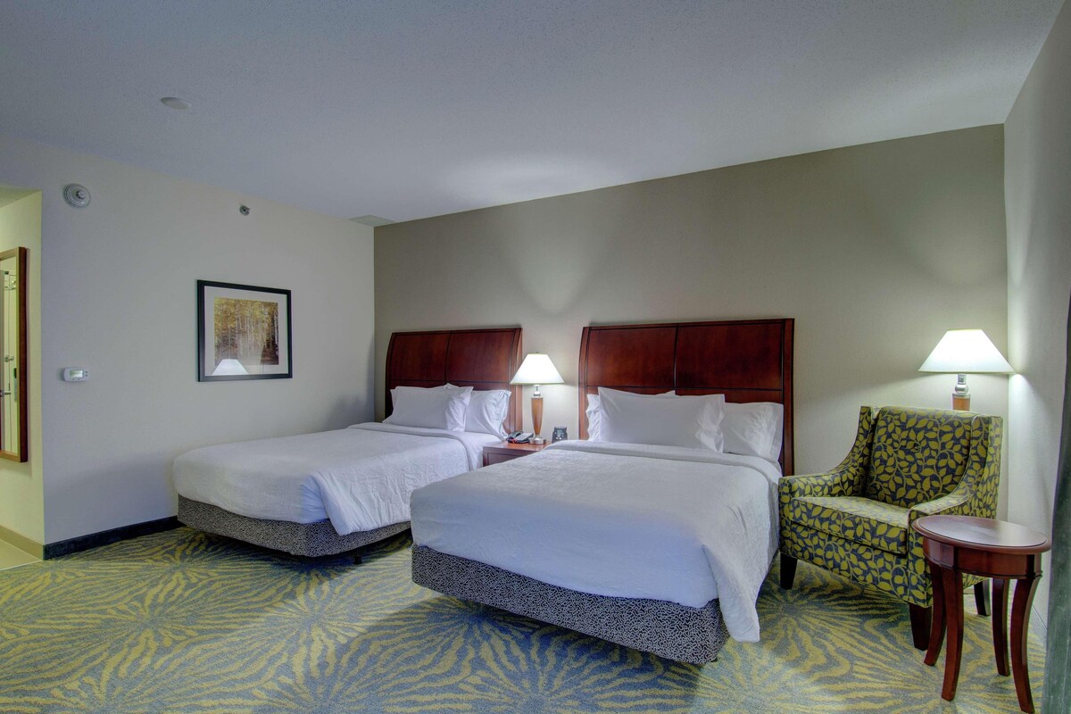 A hotel room with two beds, a chair, a small table, and a lamp.