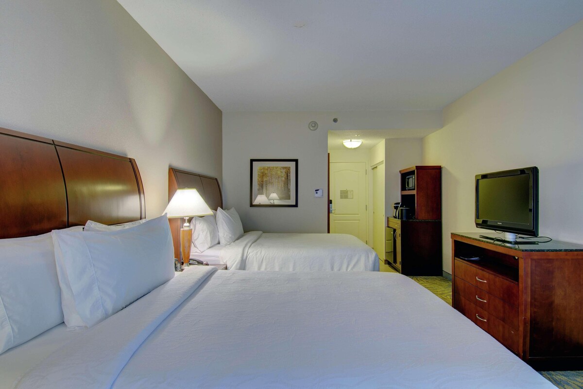 A hotel room with two beds, a TV, a lamp, and a painting on the wall.