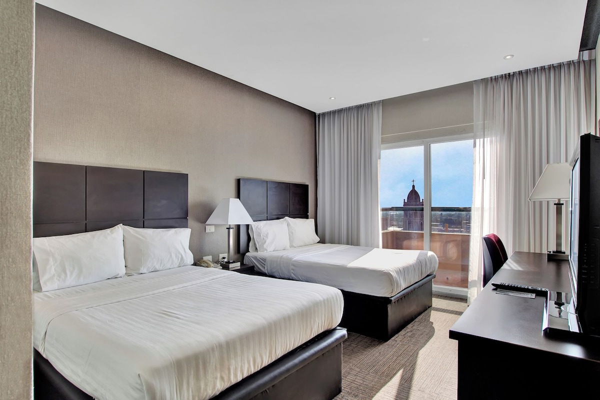 A hotel room with two beds, a desk, a chair, a TV, and a view of a cityscape.