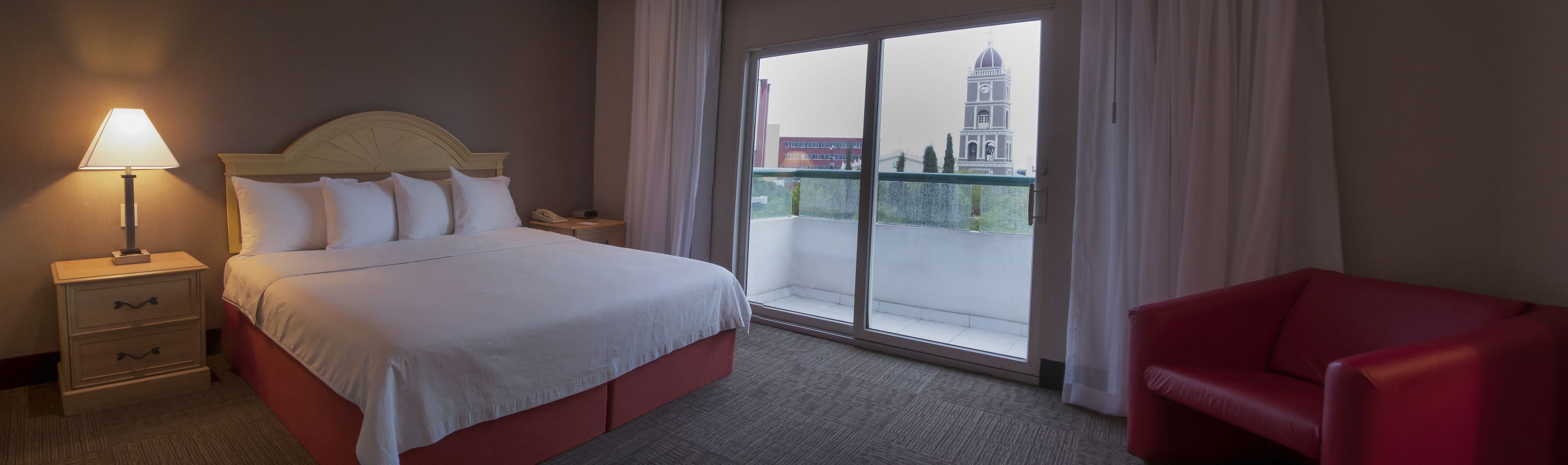 Habitación de hotel con cama, mesita de noche, una silla roja y un amplio ventanal con vista a un edificio con torre del reloj.