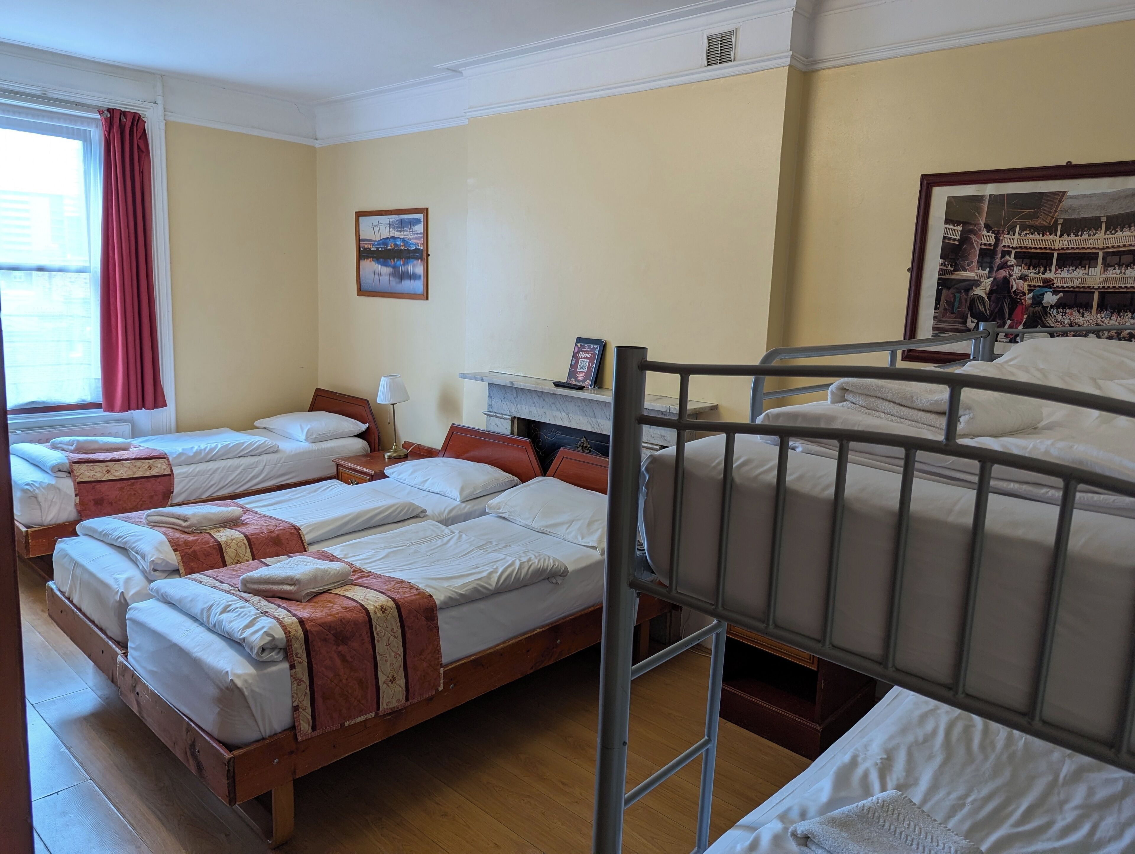 A hotel room with four beds, a fireplace, a window with curtains, and a framed picture on the wall.