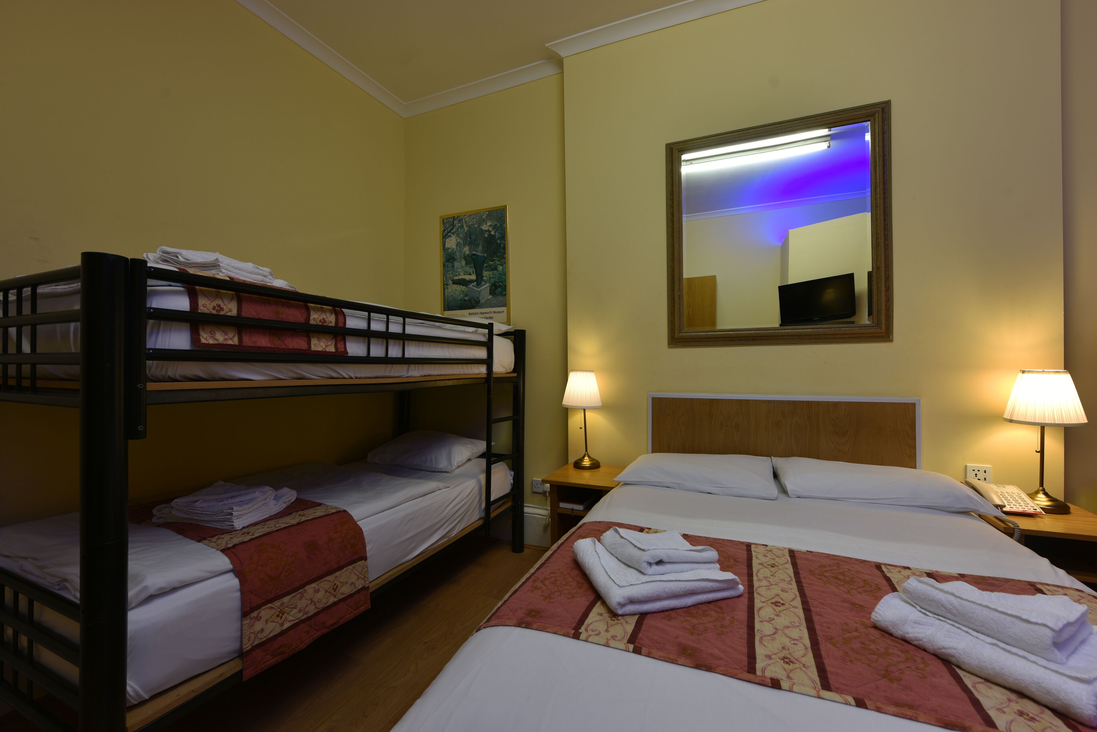 A hotel room with two beds, one with a bunk bed on top, and two bedside tables with lamps.