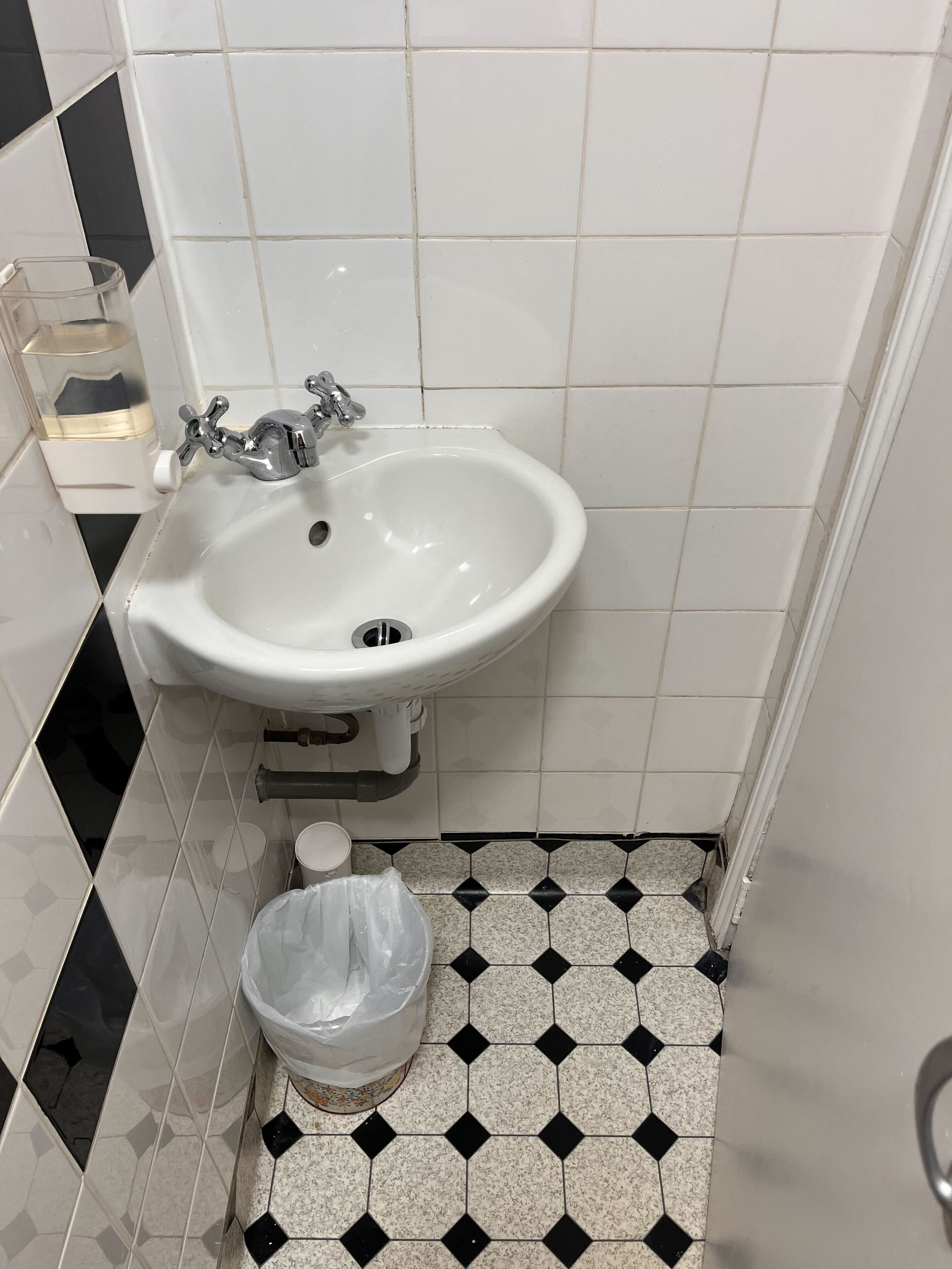 Une salle de bain avec un lavabo blanc, un sol carrelé en noir et blanc et une poubelle en plastique.