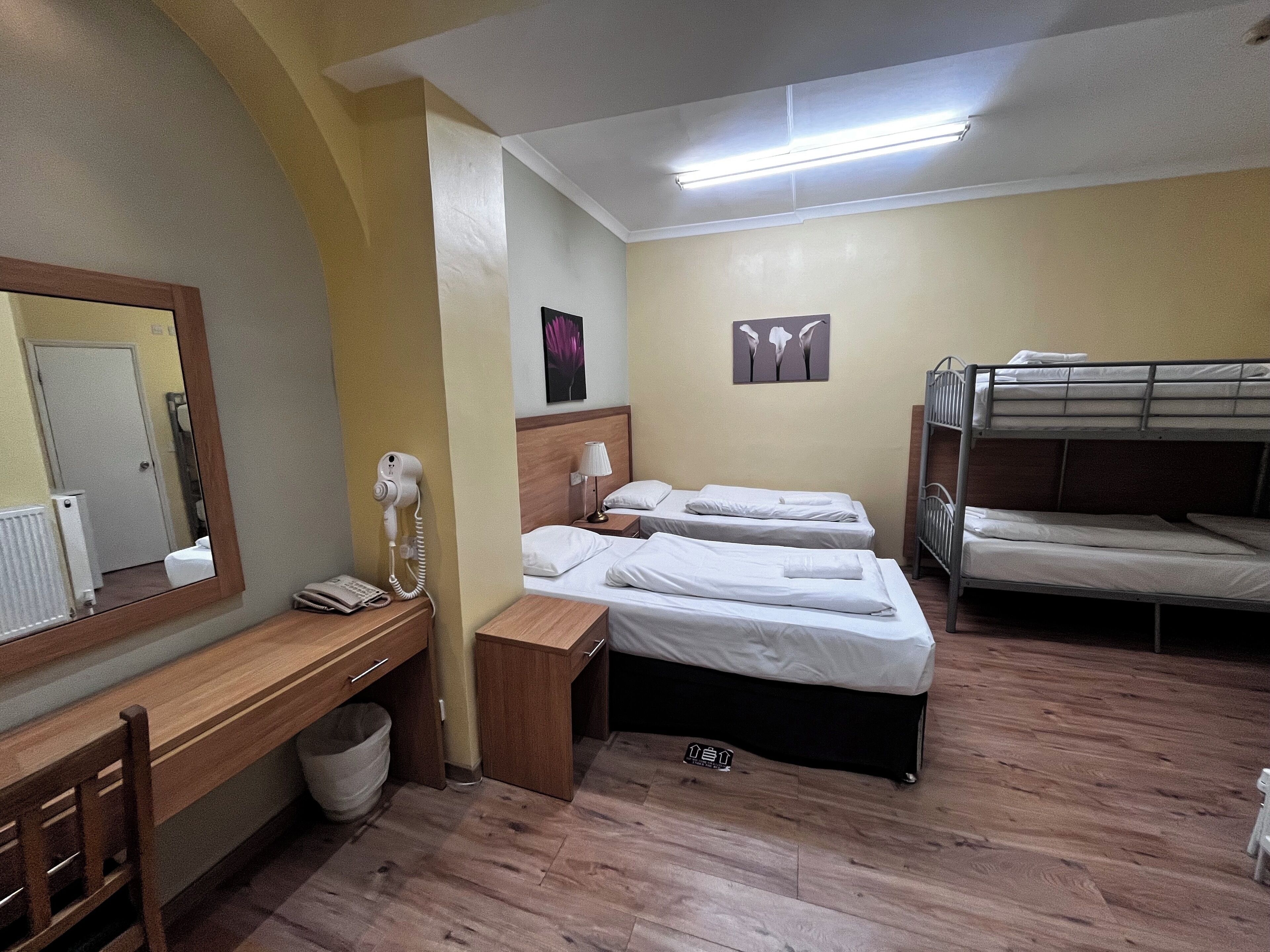 Une chambre d’hôtel avec deux lits simples, un lit superposé, un bureau avec un téléphone, un miroir et un radiateur.