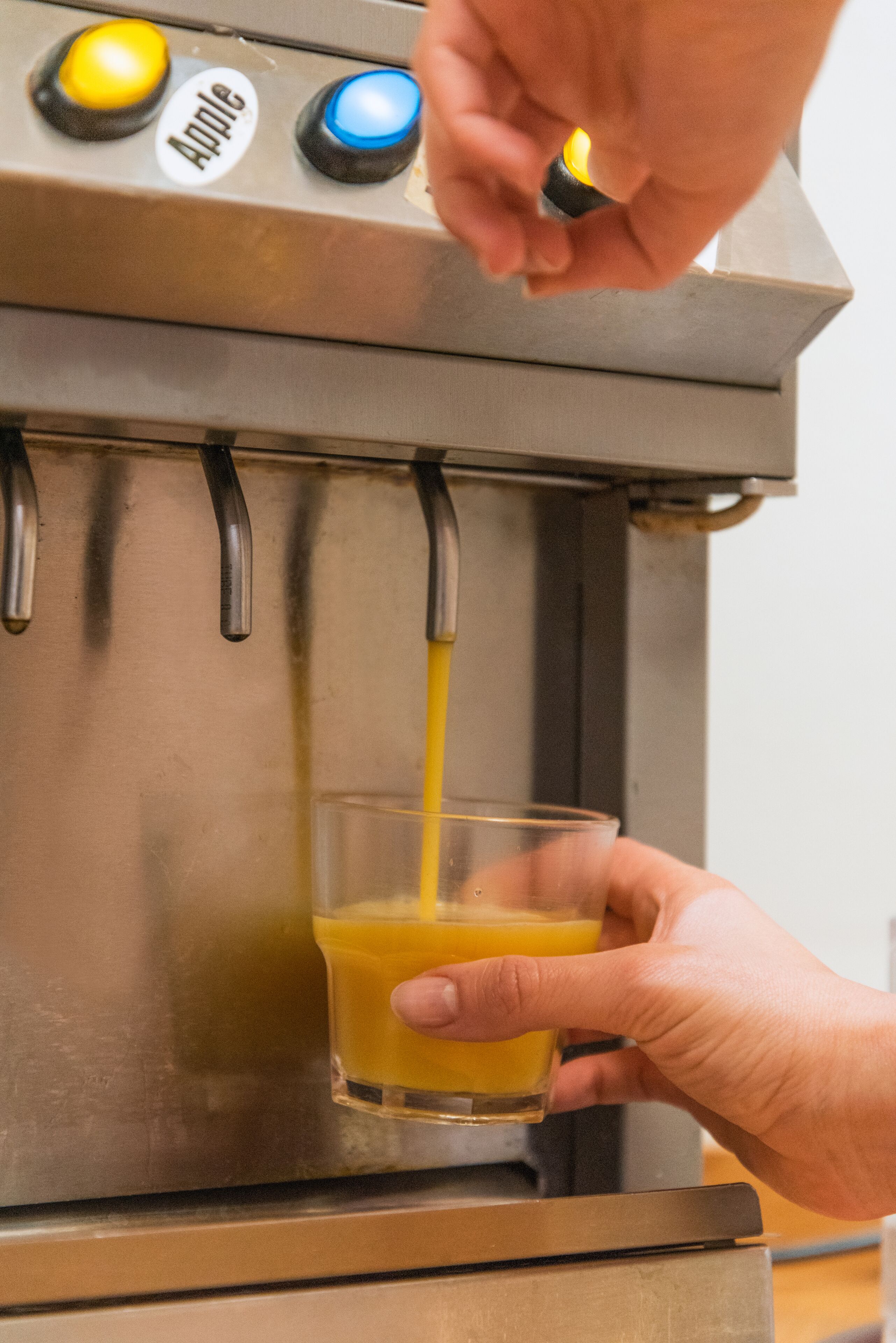 A hand holding a glass receiving orange juice from a machine with buttons labeled "Apple" and "Blue".