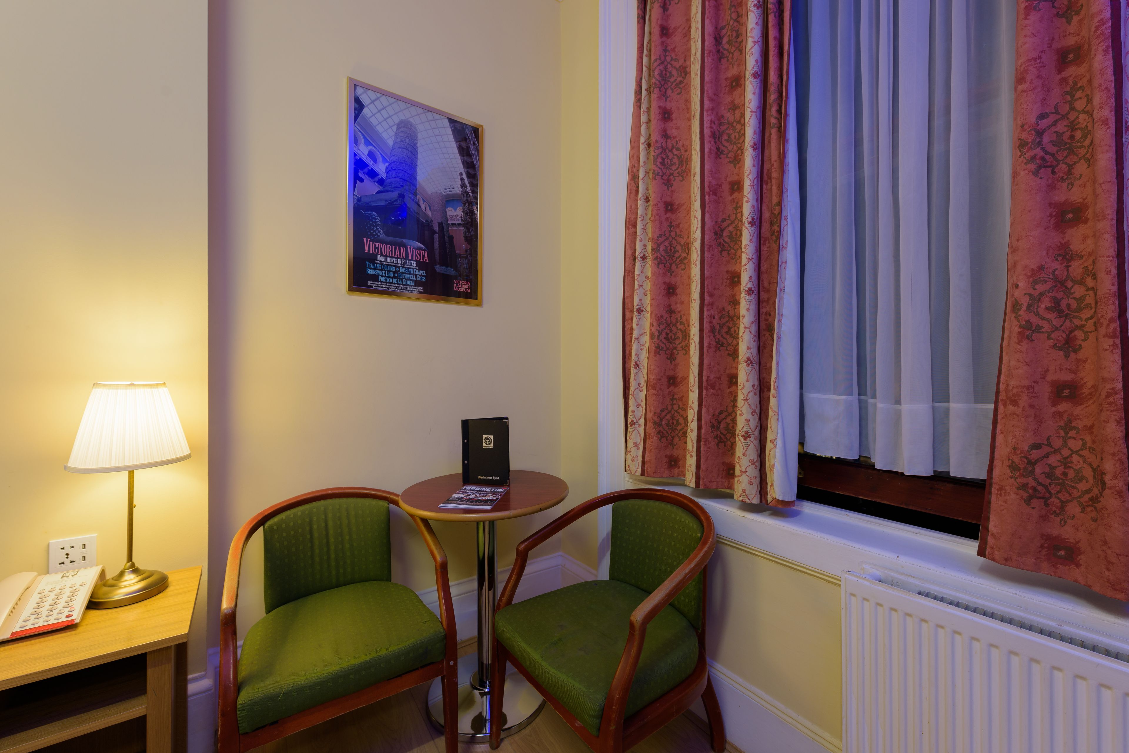 Une pièce comprenant deux fauteuils verts, une petite table ronde avec un livre, une lampe et une affiche accrochée au mur.