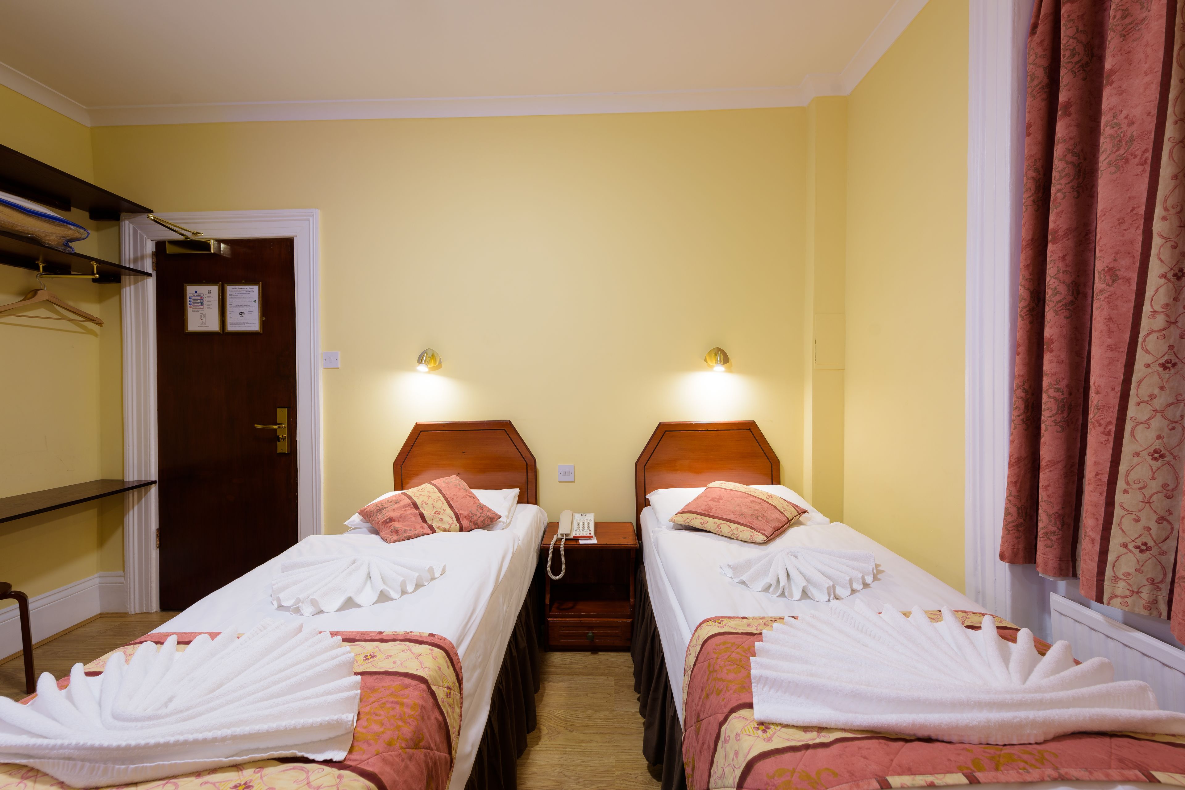 A hotel room with two single beds, a wooden nightstand with a telephone, and a door with a coat rack.