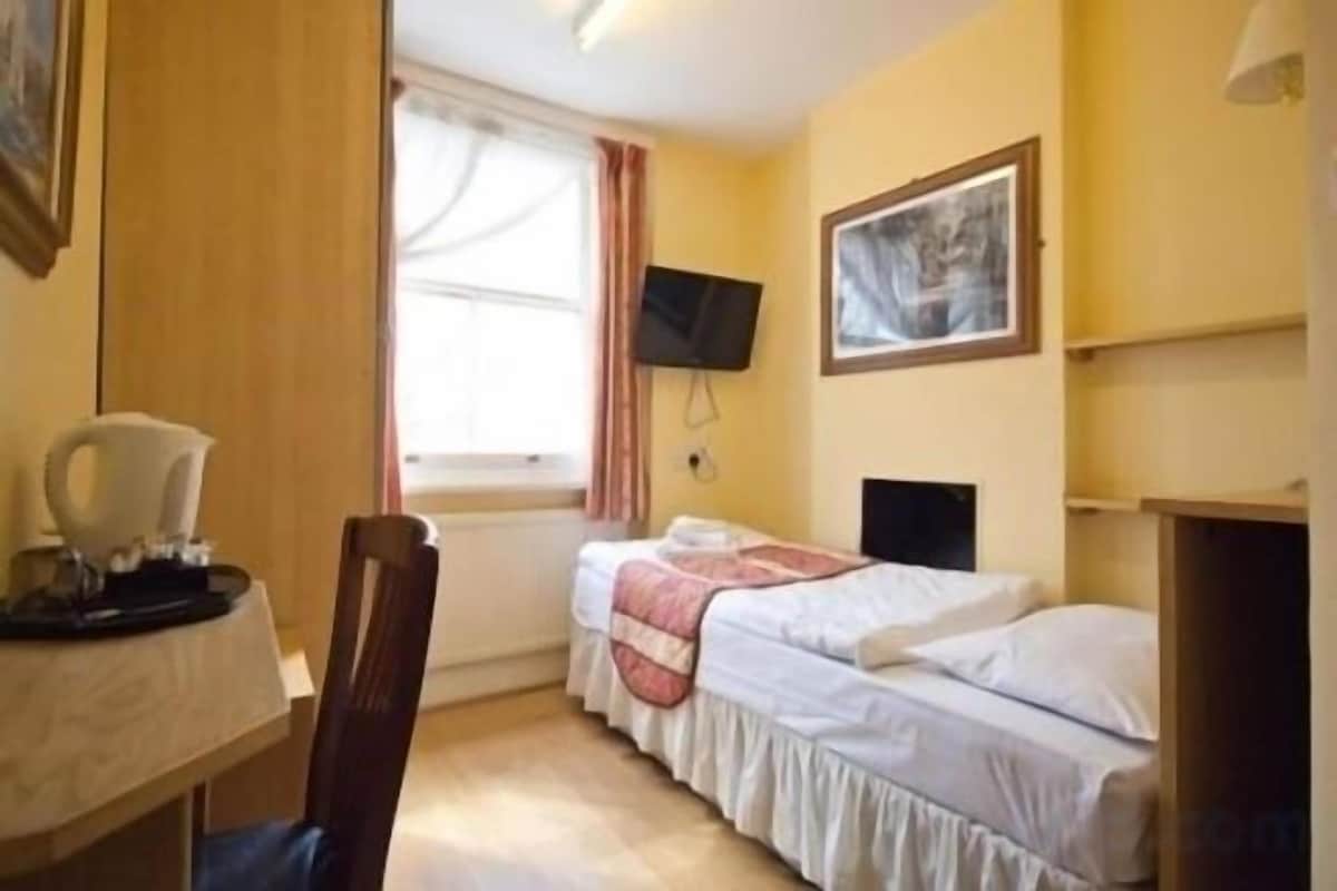 Een kleine, compacte kamer met twee eenpersoonsbedden, een bureau met stoel, een aan de muur bevestigde televisie en een raam met gordijnen.