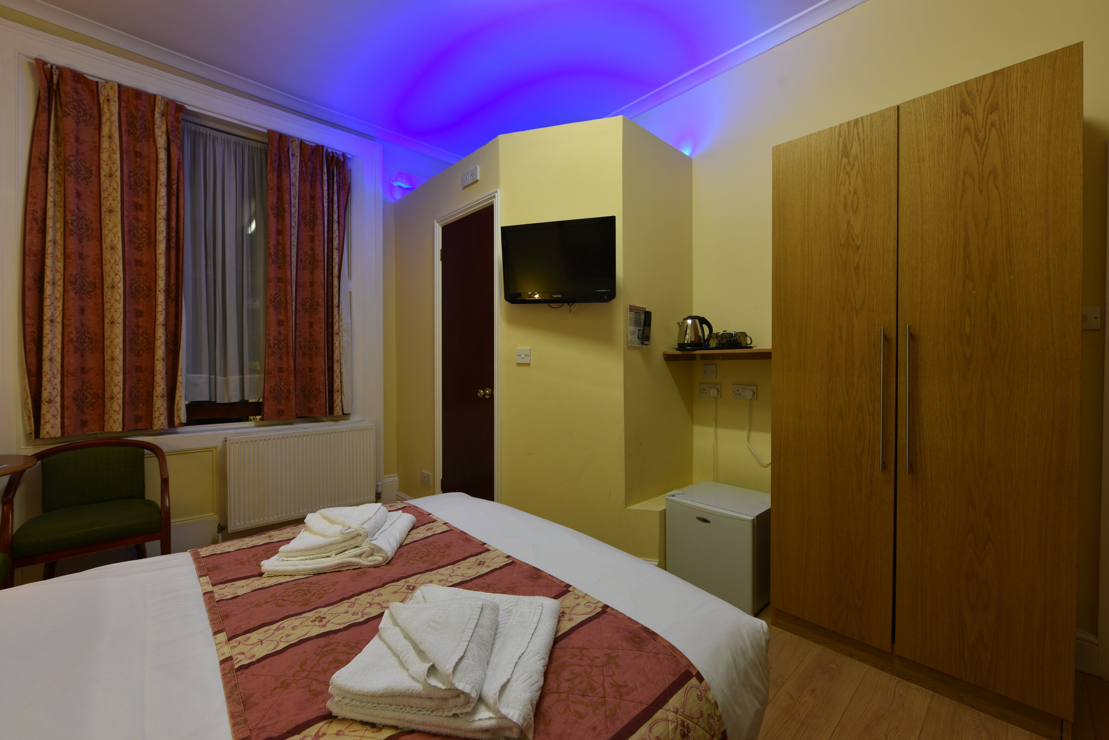 A hotel room with a bed, a chair, a wardrobe, a television, and a radiator.