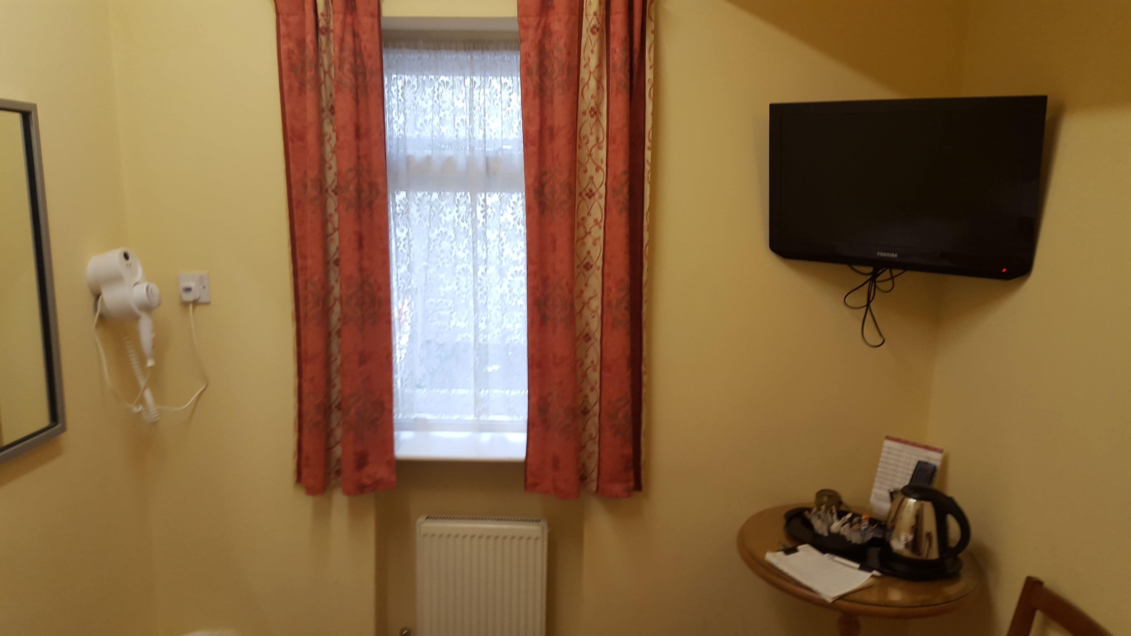 A room with a radiator, a hairdryer, a television mounted on the wall, and a small table with a kettle and papers.
