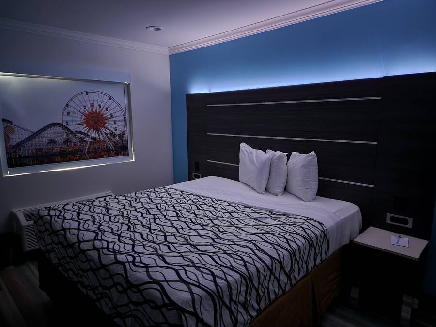 A modern hotel room with a large bed, a headboard with horizontal lines, a wall-mounted artwork of a Ferris wheel, and a bedside table with a lamp.
