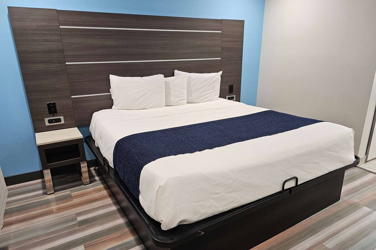 Una cama bien hecha con ropa de cama blanca y un detalle azul, una mesita de noche con un teléfono y un panel de control fijado a la pared.