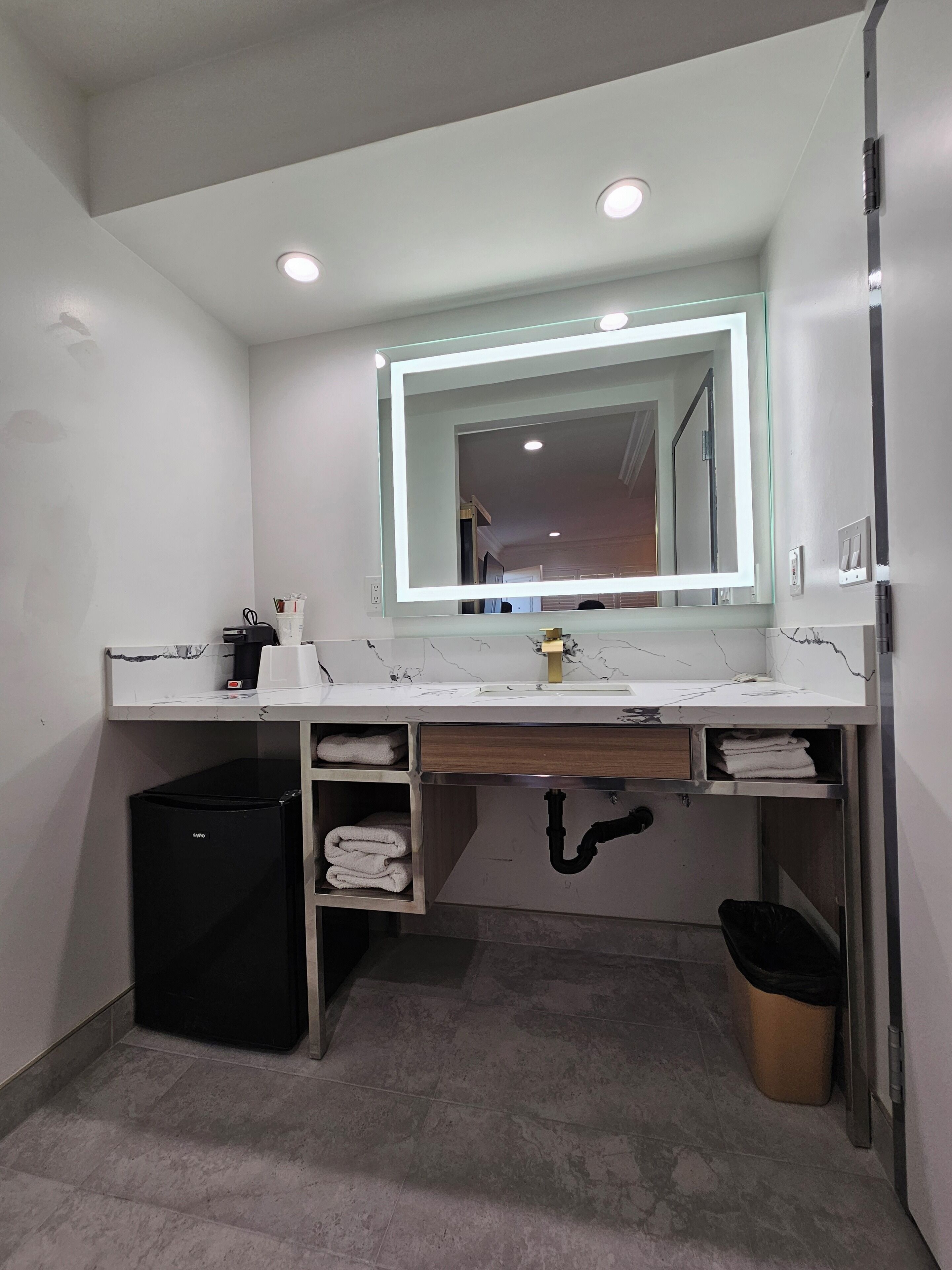 Casa de banho moderna com toucador branco, um espelho grande com iluminação LED, um cesto de lixo preto e um porta-toalhas.