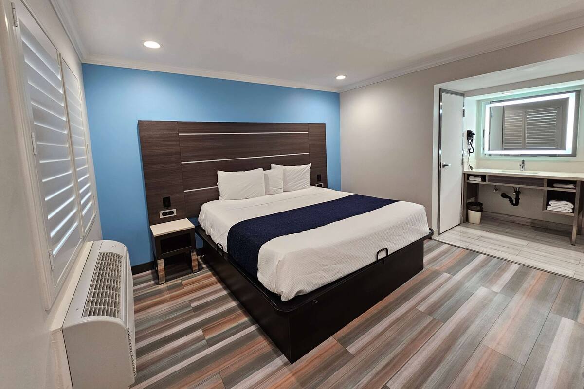 Una habitación de hotel moderna con una cama grande, una mesita de noche, un armario empotrado y un baño con espejo y lavamanos.