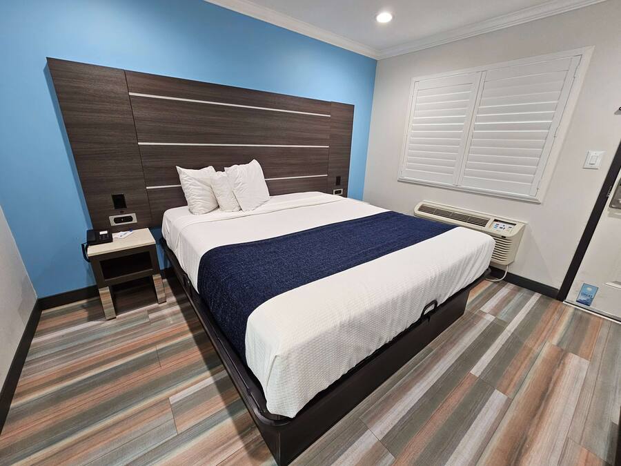 A neatly made bed with white and blue bedding, a bedside table with a phone and papers, a wall-mounted air conditioner, and striped wooden flooring.