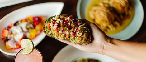 A hand holding a slice of toast topped with various ingredients, a bowl of salad, and a plate of food in the background.