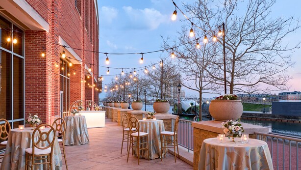 Un patio al aire libre con mesas y sillas, luces colgantes y macetas grandes.