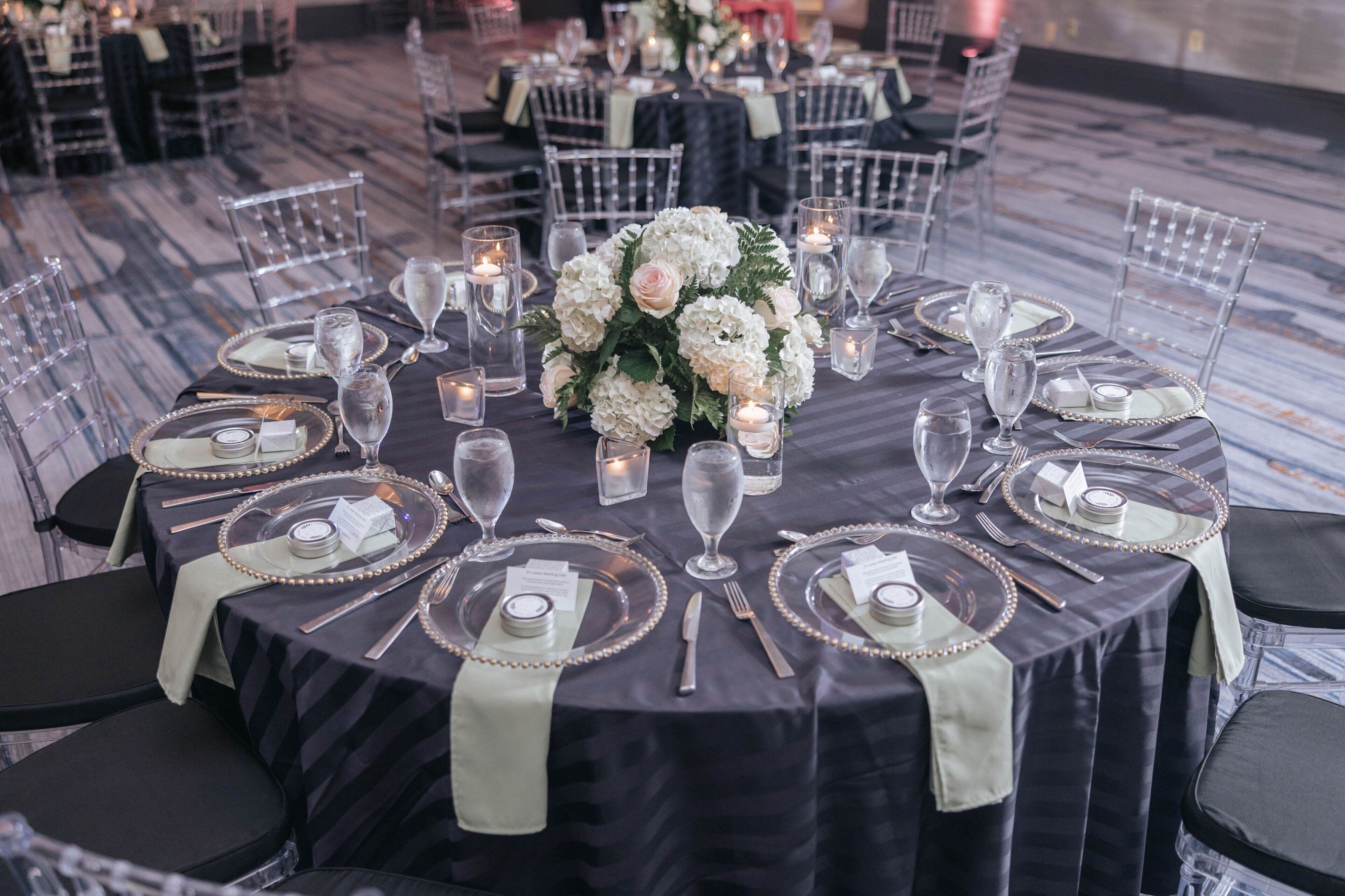 A round table set for a formal event with clear chairs, a dark tablecloth, and a centerpiece of white flowers.