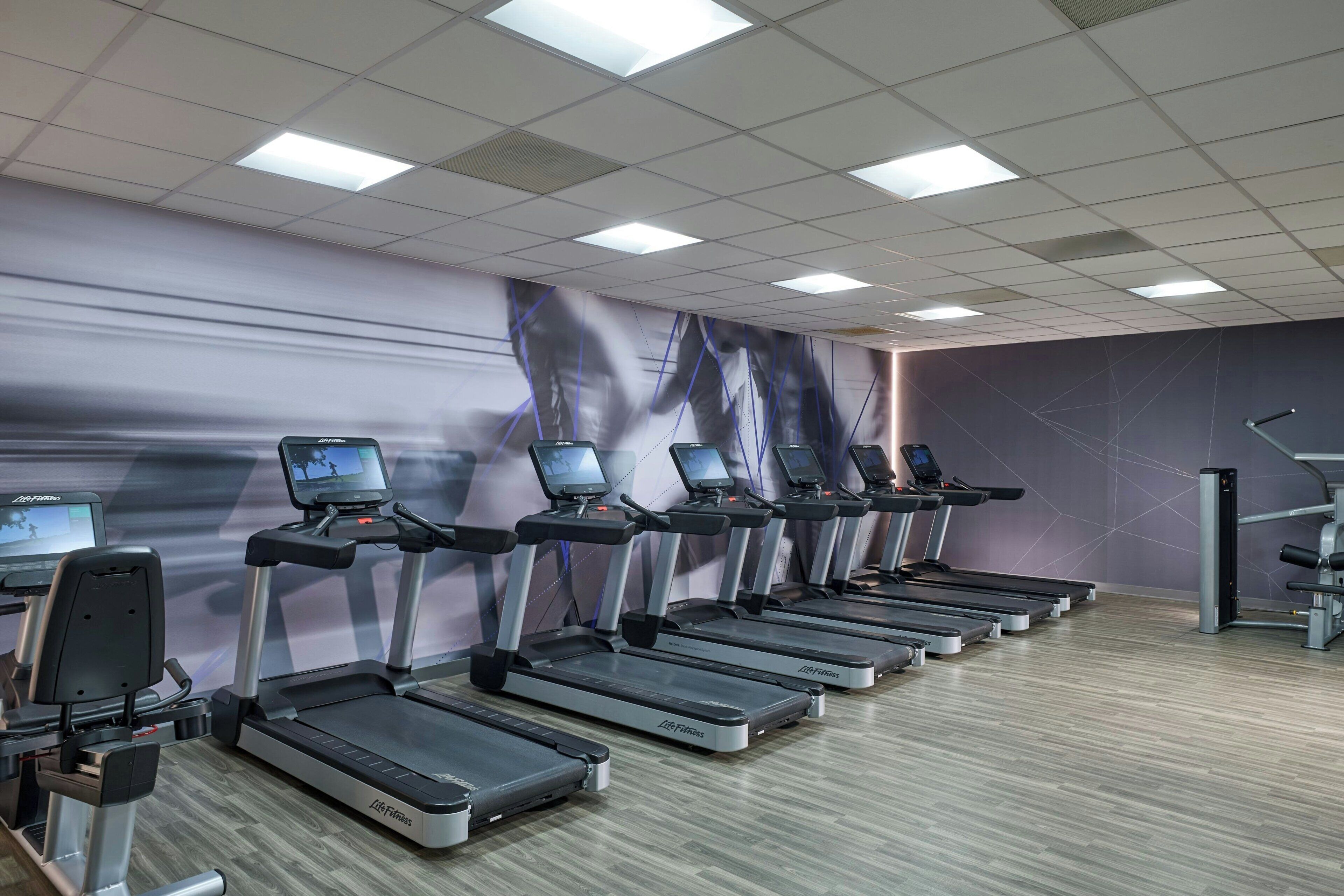 Une salle de sport équipée de plusieurs tapis de course, d’appareils d’exercice et d’un mur orné d’une grande affiche.