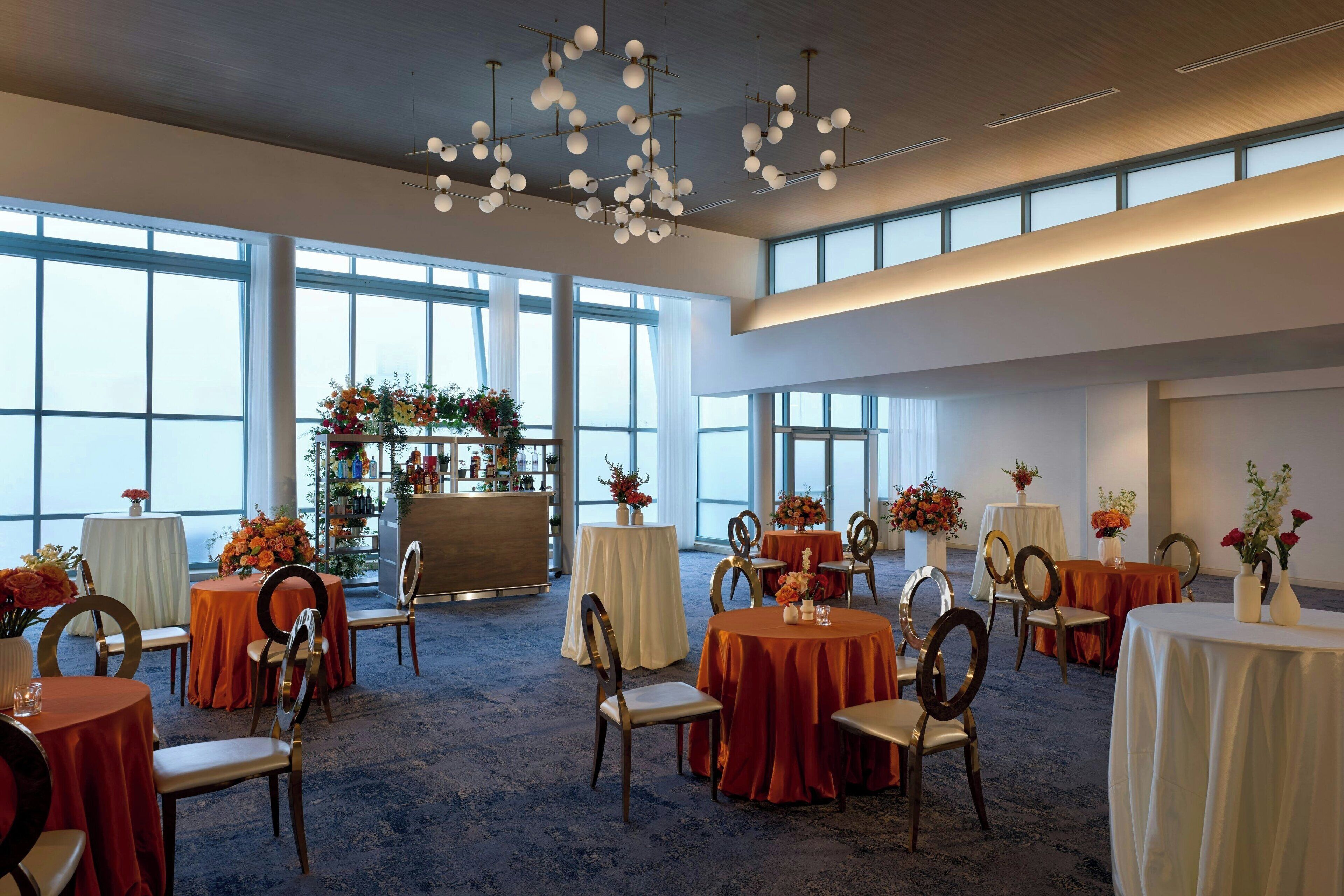 A spacious conference room with round tables, chairs, and a chandelier. Large windows provide natural light.