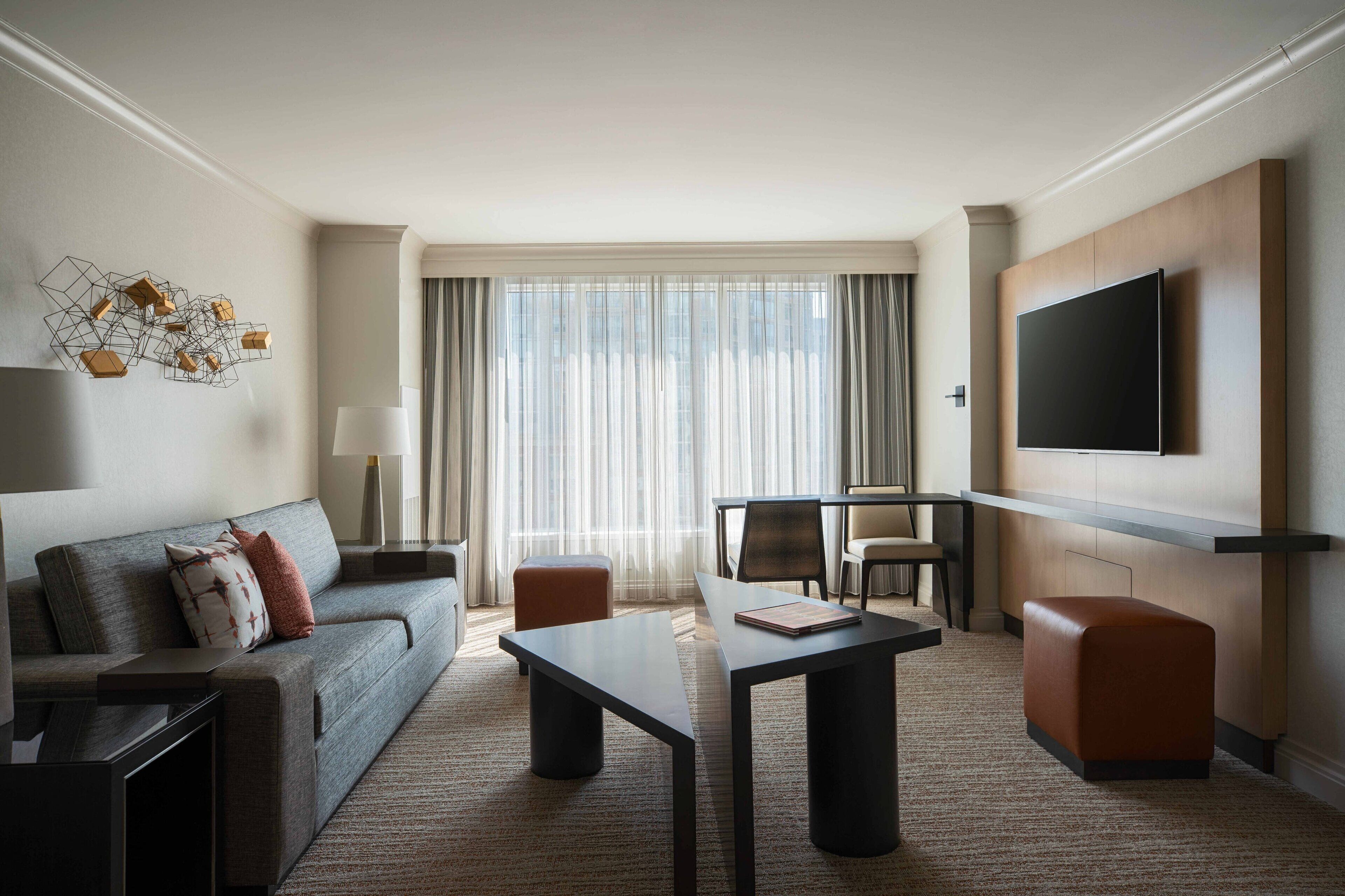 Una habitación de hotel moderna con sofá, una mesa de comedor, televisión montada en la pared y un ventanal con cortinas translúcidas.