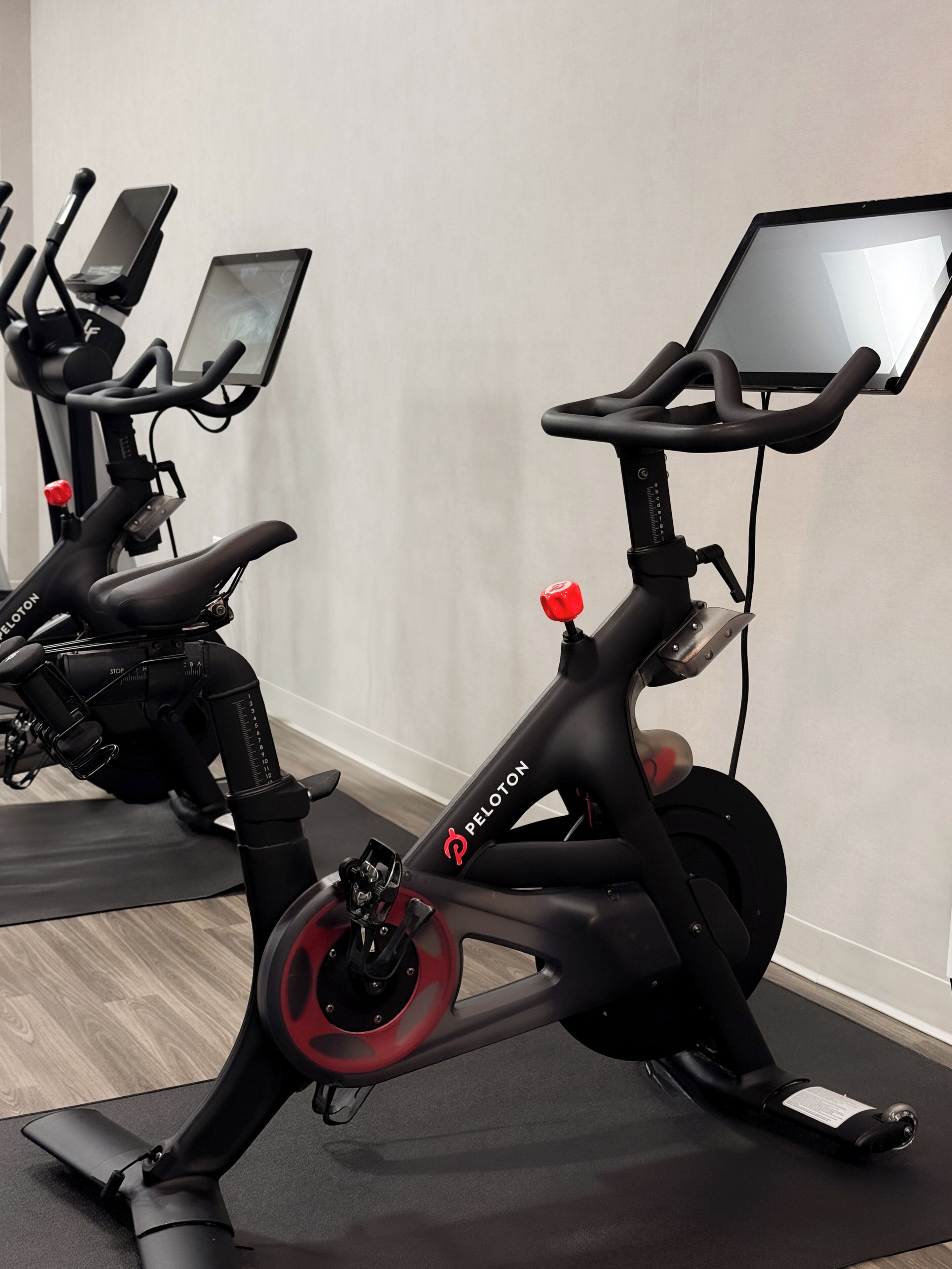 A stationary bike with a digital display and a red knob, positioned on a mat in a room with a plain wall.