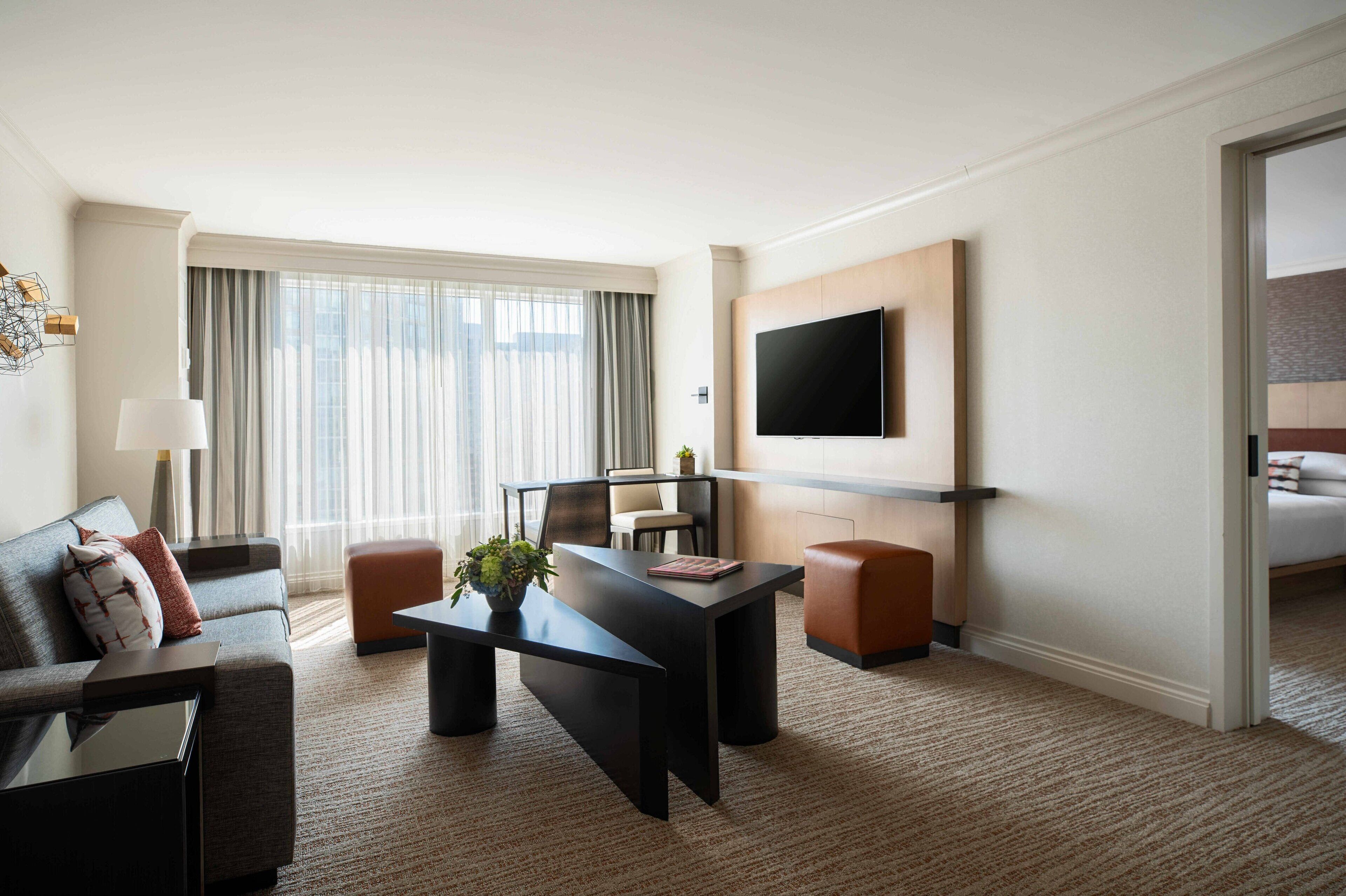 A modern hotel room with a sofa, a dining table, a TV mounted on the wall, and a large window with curtains.