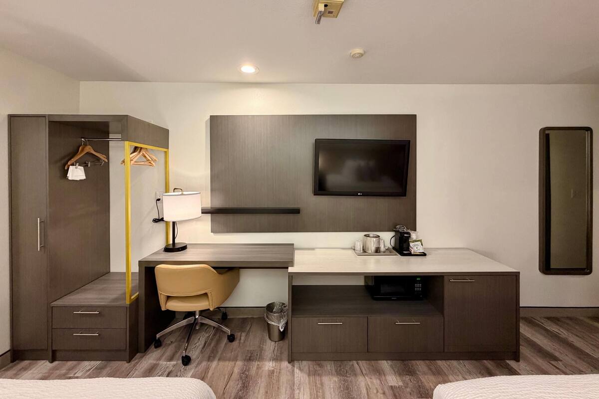A modern hotel room with a desk, chair, TV, wardrobe, and a small kitchenette.