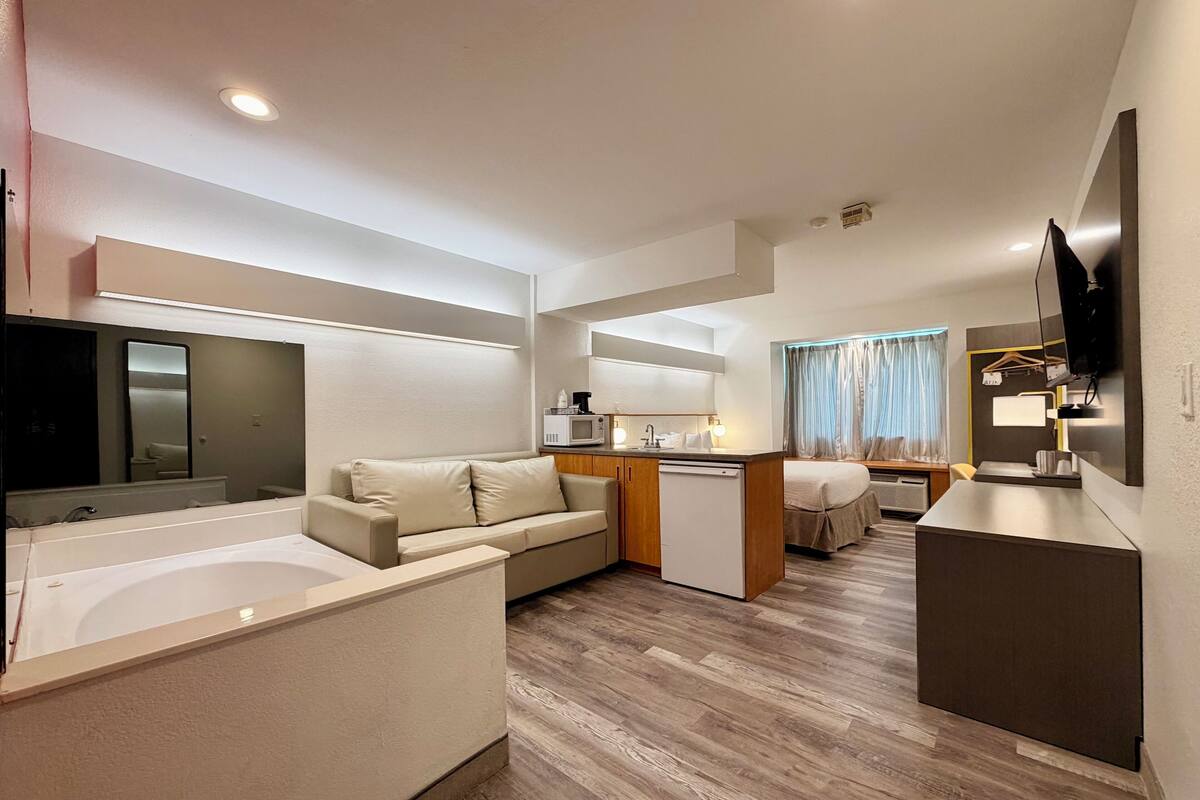 A modern hotel room with a kitchenette, a sofa, a small dining area, a TV, and a large window with curtains.