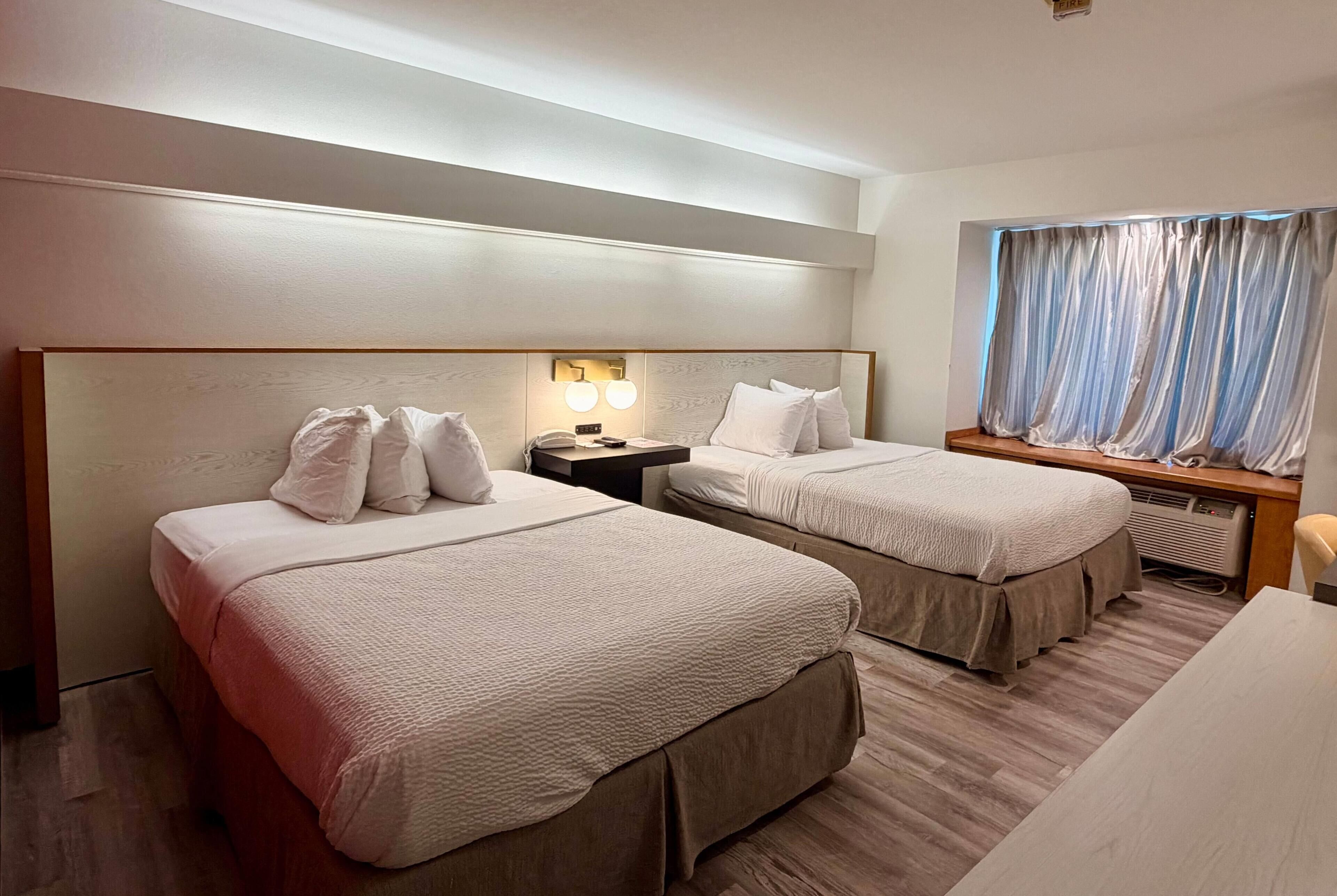 A hotel room with two beds, a nightstand, a desk, and a window with curtains.