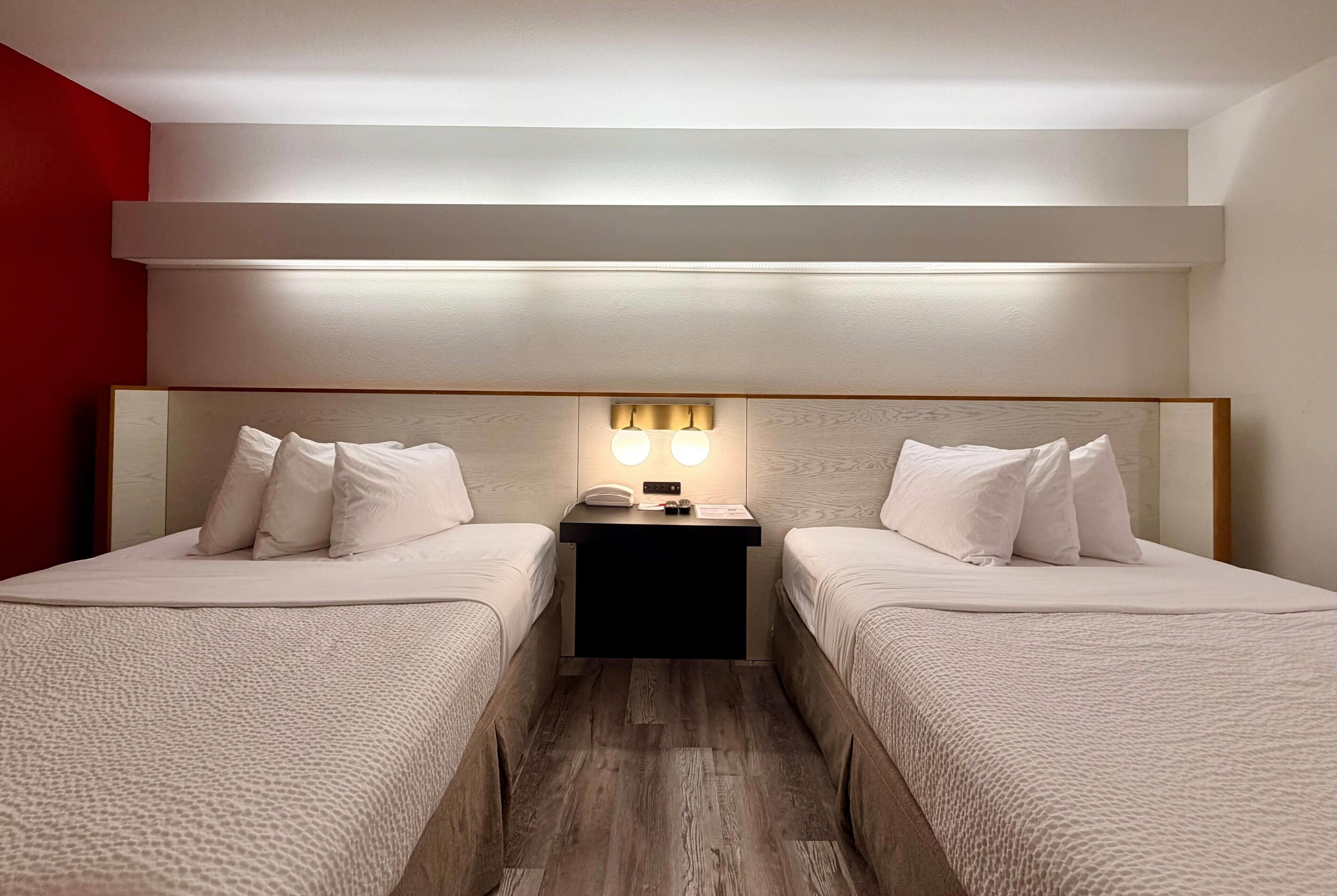 A hotel room with two beds, a bedside table with a lamp, and a red accent wall.