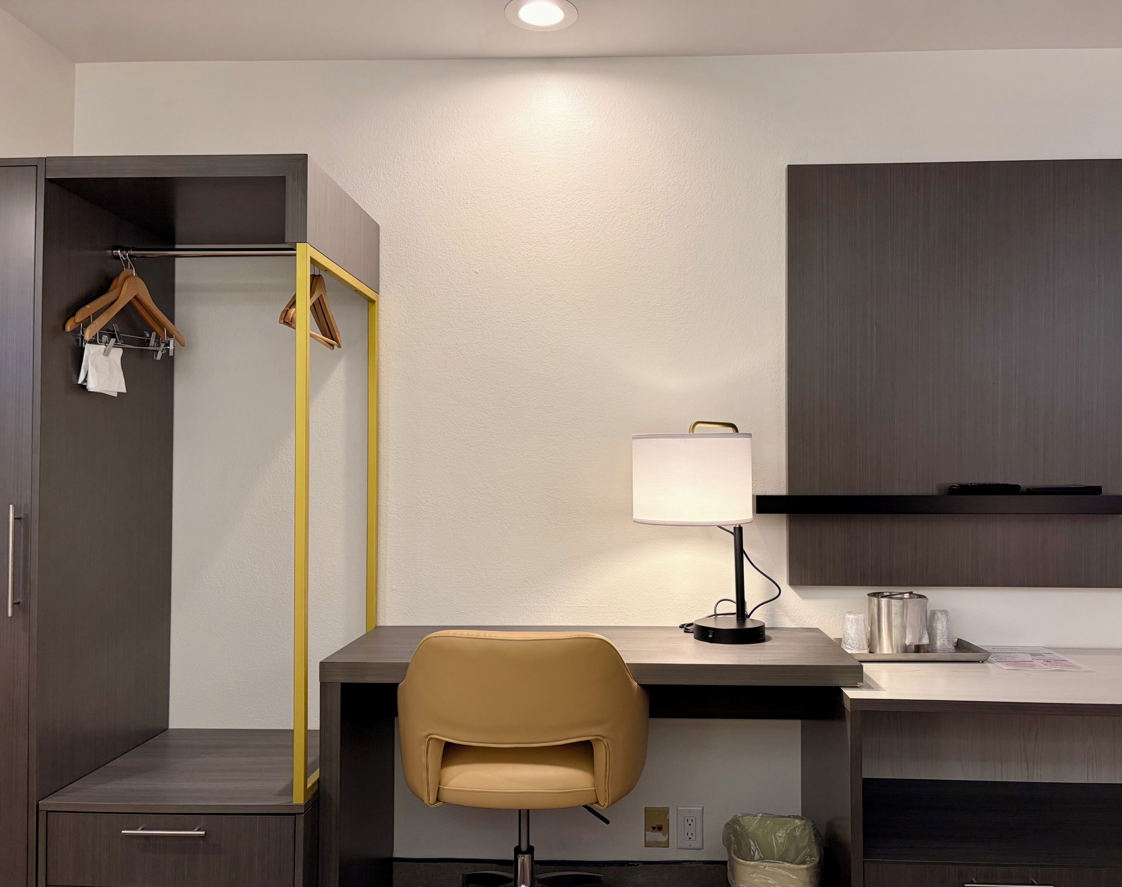 A hotel room with a desk, chair, wardrobe, hanging clothes rack, lamp, and a trash bin.