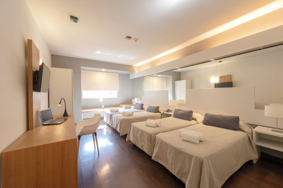 A hotel room with four single beds, a desk with a laptop, a chair, and a large window.