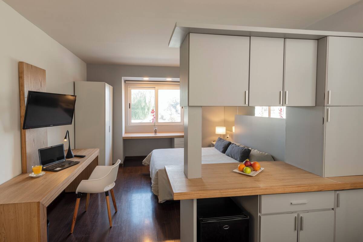 A compact hotel room with a wooden desk, a bed, a small kitchenette, and a window providing natural light.