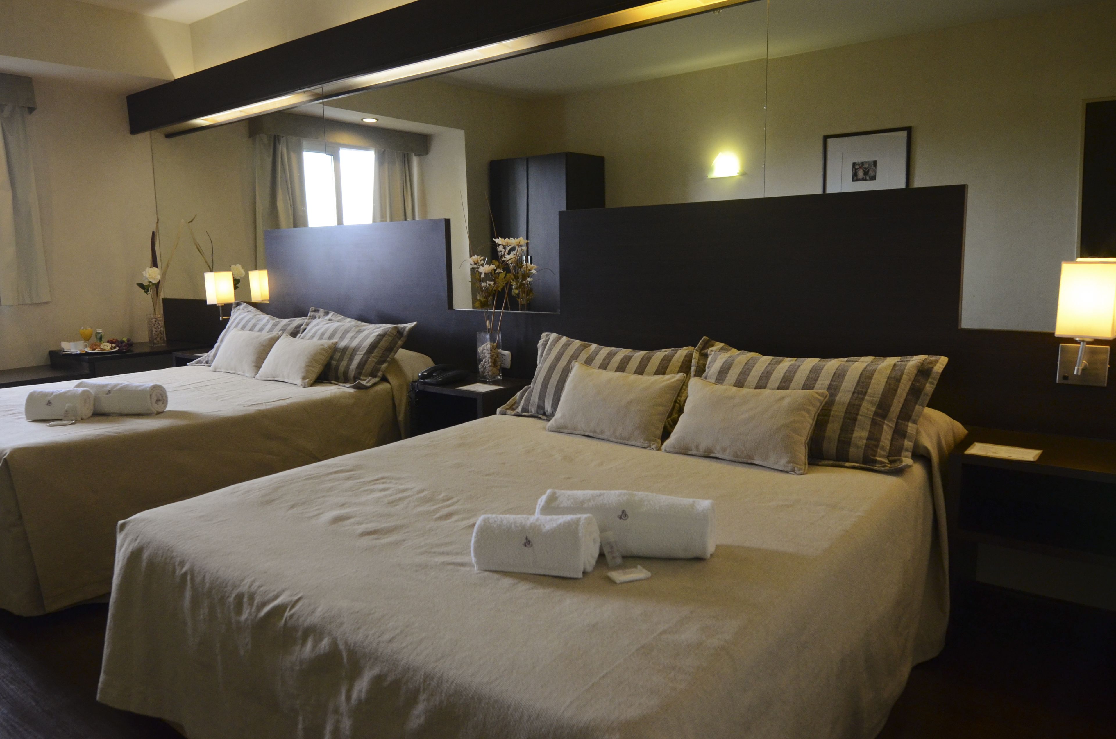 Habitación de hotel con dos camas, cada una con una almohada y una toalla enrollada. Hay una mesita de noche con una lámpara, un espejo y un florero con flores.
