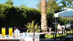 Un espace extérieur aménagé pour les repas, avec une piscine, des parasols et une table dressée avec de la nourriture et des boissons.