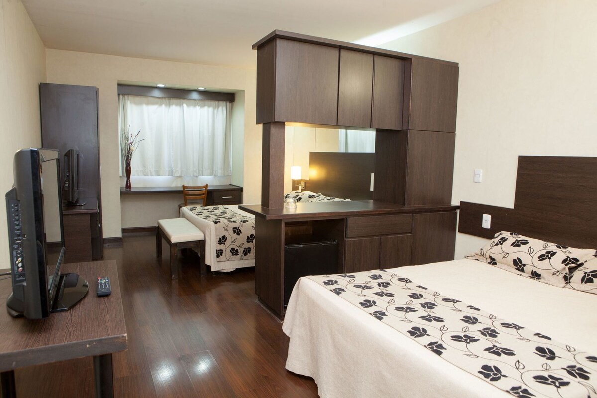 A hotel room with a bed, a desk with a chair, a television, and a window with curtains.