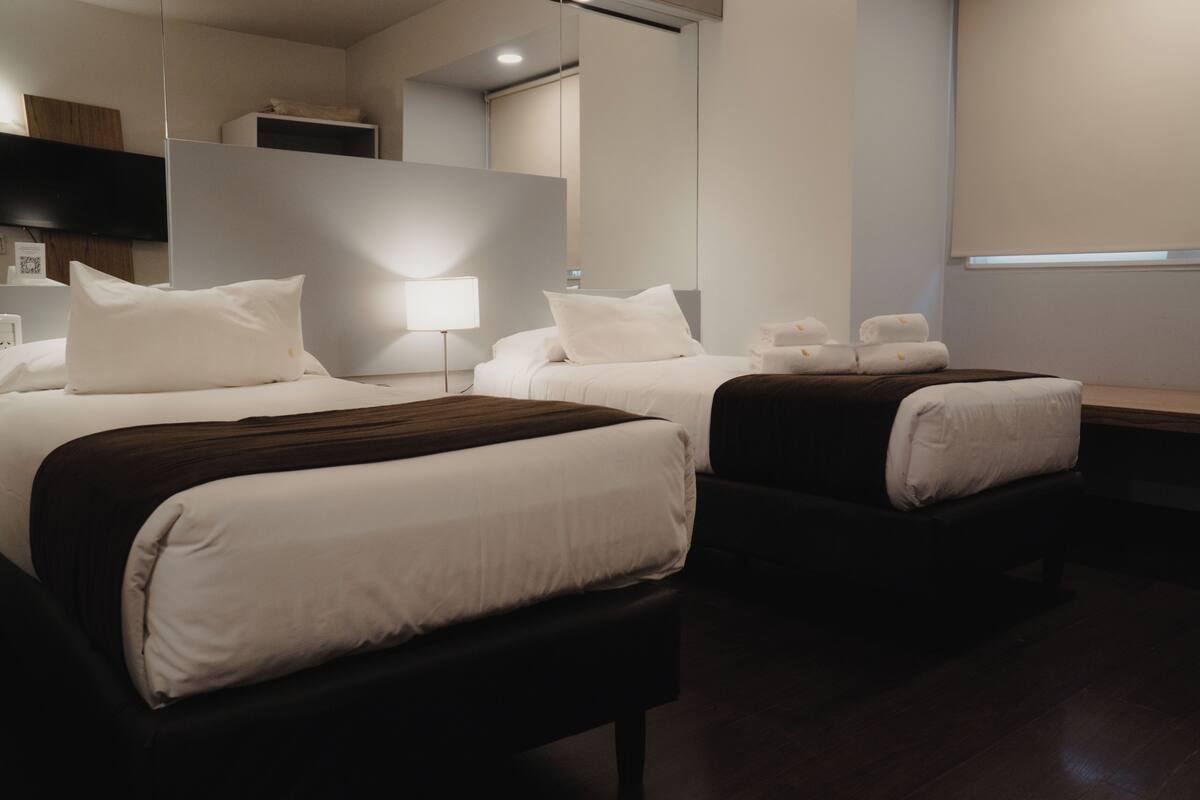 A hotel room with two beds, a bedside table with a lamp, a TV mounted on the wall, and a window with blinds.