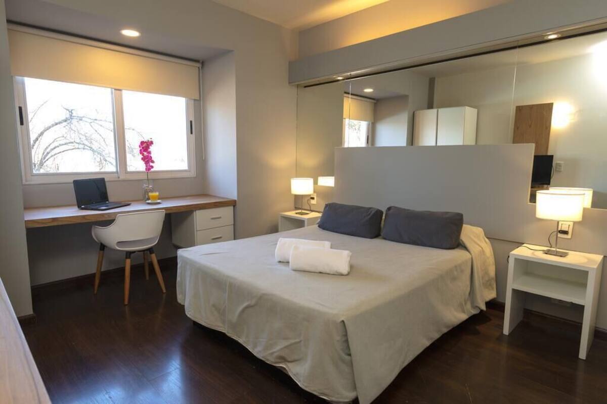 A modern hotel room with a large bed, a desk, and a chair. There is a window with a view of trees and a lamp on each side of the bed.