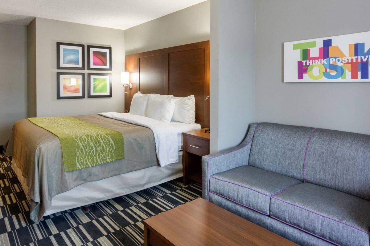 A hotel room with a bed, a gray sofa, a wooden nightstand, and a wall with framed pictures and a motivational poster.